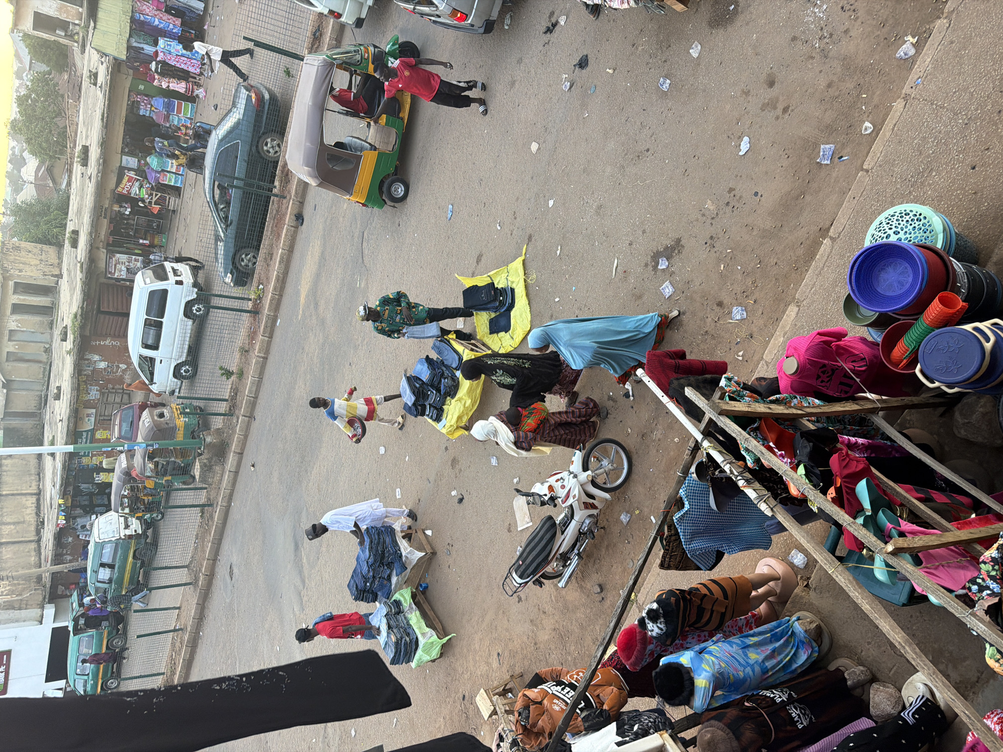 This image captures a bustling street market scene in Bukuru, Nigeria. A busy road runs through the center, lined with shops and vendors. Various vehicles, including vans and a motorized rickshaw, are present on the road. Several individuals are engaged in commercial activities, with piles of clothing, primarily jeans, displayed for sale on tarps and tables. Some people are interacting with vendors, while others are seen walking or standing around. The environment appears to be an outdoor urban setting with a mix of built structures and open ground. The overall impression is one of a vibrant marketplace with economic activity and community interaction.