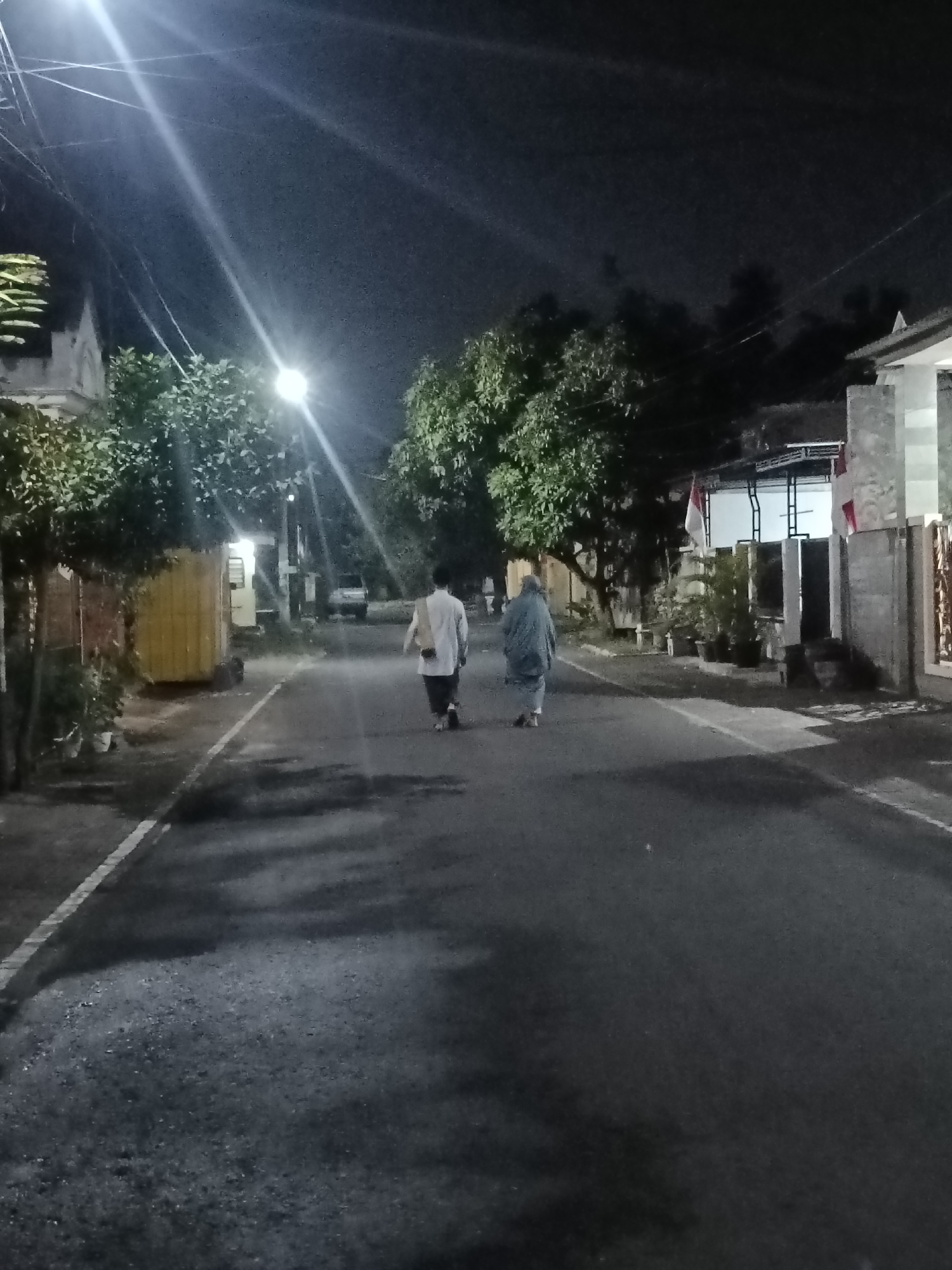 At night on a paved street in Kediri City, Indonesia, two individuals walk away from the camera. The person in front is wearing a long, light-colored garment, possibly a shirt or tunic, over dark pants. They have a light-colored bag or scarf draped over their left shoulder. The person behind them is wearing a long, loose, dark-colored garment, consistent with a hijab or abaya. They are walking in the same direction. The street is illuminated by a bright street lamp overhead, casting strong light and shadows on the asphalt. Trees line the sides of the street, with some foliage visible. Buildings with visible windows and doorways are present on both sides of the street. A white line marks the edge of the road on the left. In the distance, a vehicle is parked on the left side of the road. The overall scene depicts a quiet, residential street at night.