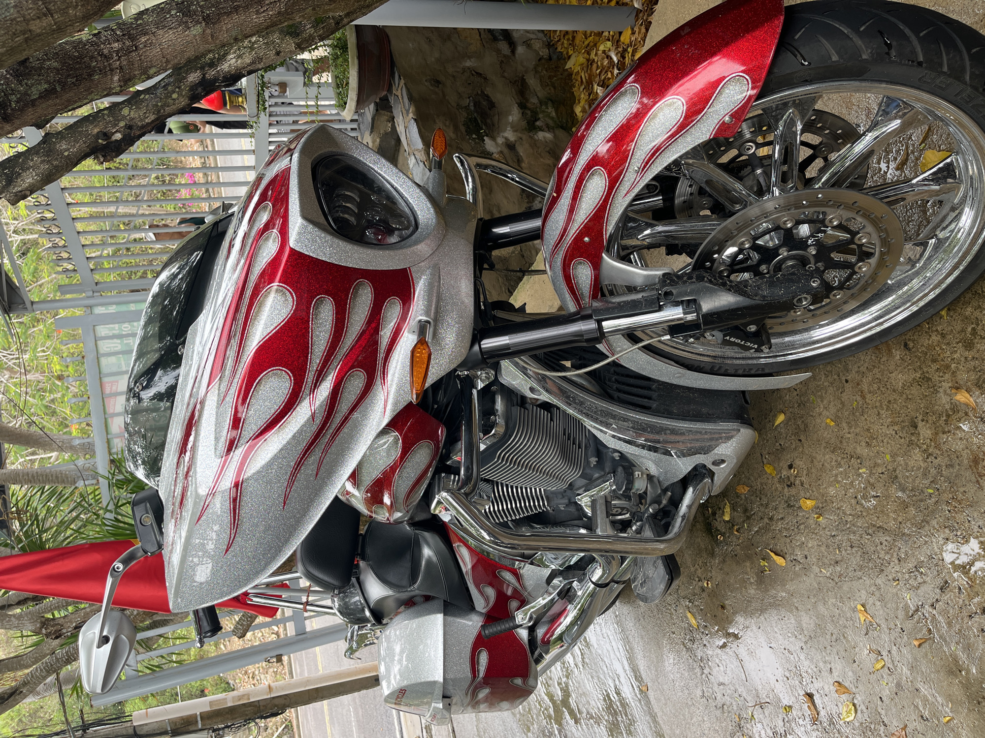 A silver and red Victory motorcycle with flame graphics is parked on a wet concrete surface. The location appears to be outdoors, possibly near a residence with a gate. The motorcycle is positioned at an angle, showcasing the front wheel and engine. Leaves are scattered on the ground. The photograph was taken in or around Bac Nha Trang Ward, Vietnam.