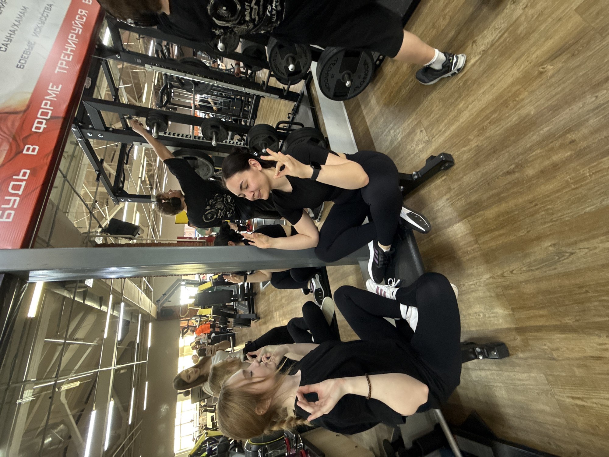 A group of four young women are posed in a gym with a focus on capturing a photo or video, likely for social media purposes. The women are all dressed in black workout attire, indicating a synchronized theme or exercise routine.
The gym setting is typical, with weightlifting equipment and other fitness apparatus visible in the background. A banner or poster displaying Russian text that includes the words "Be in shape Train" can be seen, indicating that the location is likely in a Russian-speaking area.
The photograph appears to have been taken indoors, with artificial lighting evident from the overhead fixtures. The composition suggests that the photographer is positioned at a slight angle to the subjects, possibly using a wide-angle lens to capture the entire group within the frame.