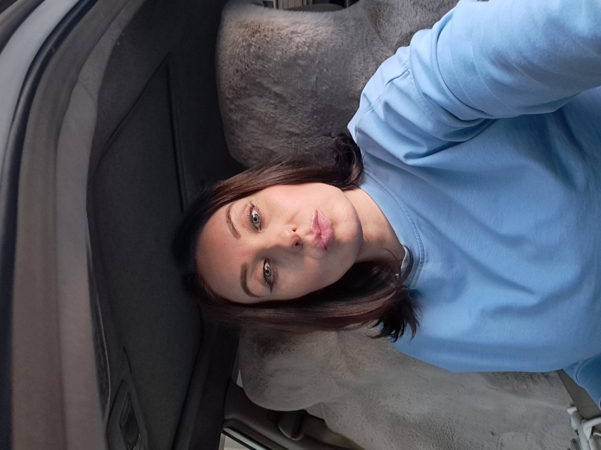 A woman is captured in a selfie taken from inside a vehicle. She is positioned in the driver's seat, with the car's interior visible in the background. The car's headliner, a sunroof, and the back of the passenger seat are discernible. The woman has medium-length brown hair and is wearing a light blue long-sleeved shirt. She is looking directly at the camera with her lips pursed and appears to be wearing makeup, including eyeliner and lipstick. The lighting suggests it may be daytime. The specific location within the city of Irkutsk, Russia, cannot be determined from the image alone.