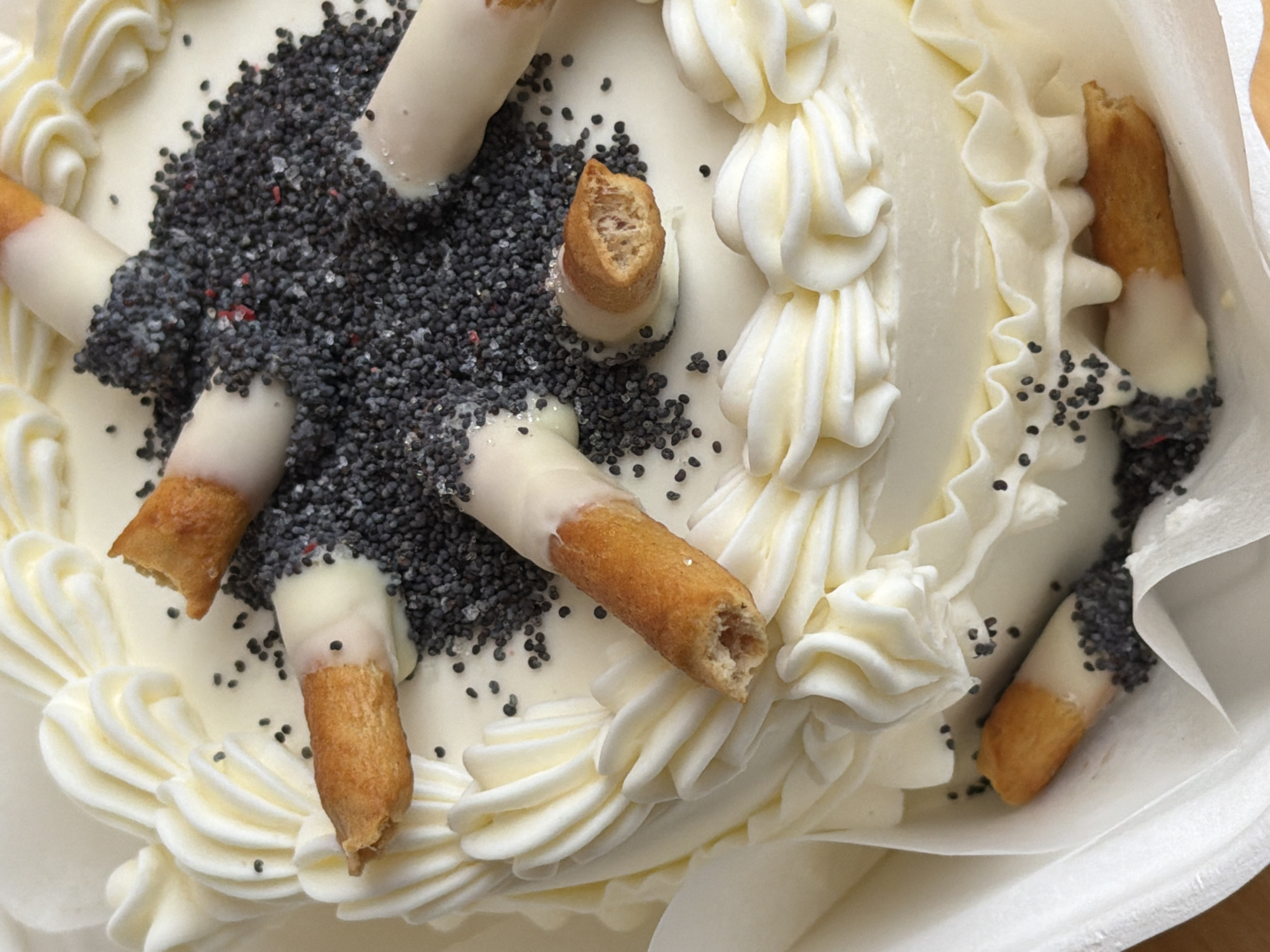 The image depicts a close-up, overhead shot of a cake. The cake is decorated with white frosting, piped in a swirl pattern around the edges and in decorative rosettes on top. Several cigar-shaped pastries, partially dipped in white icing and then coated in poppy seeds, are arranged across the cake's surface. A cluster of poppy seeds also appears in the center of the cake. The cake is situated within a white container. There is no information available about the location or the city of Tel-Aviv, Israel. There are no people visible.