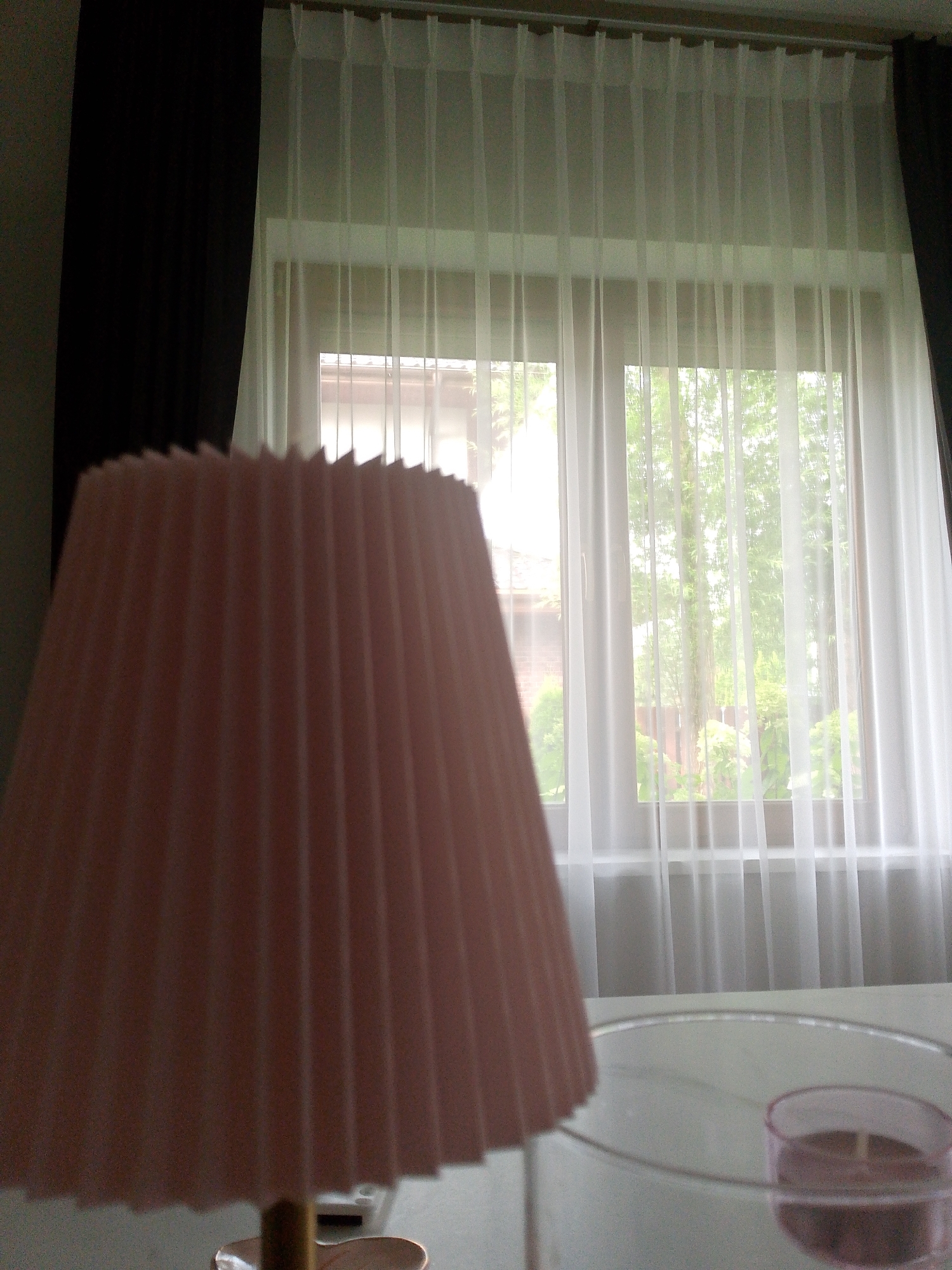 An interior shot focuses on a pleated pink lampshade in the foreground. Behind the lampshade, sheer white curtains cover a window, through which trees and greenery are visible, suggesting an outdoor environment. To the left of the window, a dark curtain hangs partially closed. On a surface below the window, a clear glass object with a pink candle inside is partially visible. The overall lighting suggests daytime.