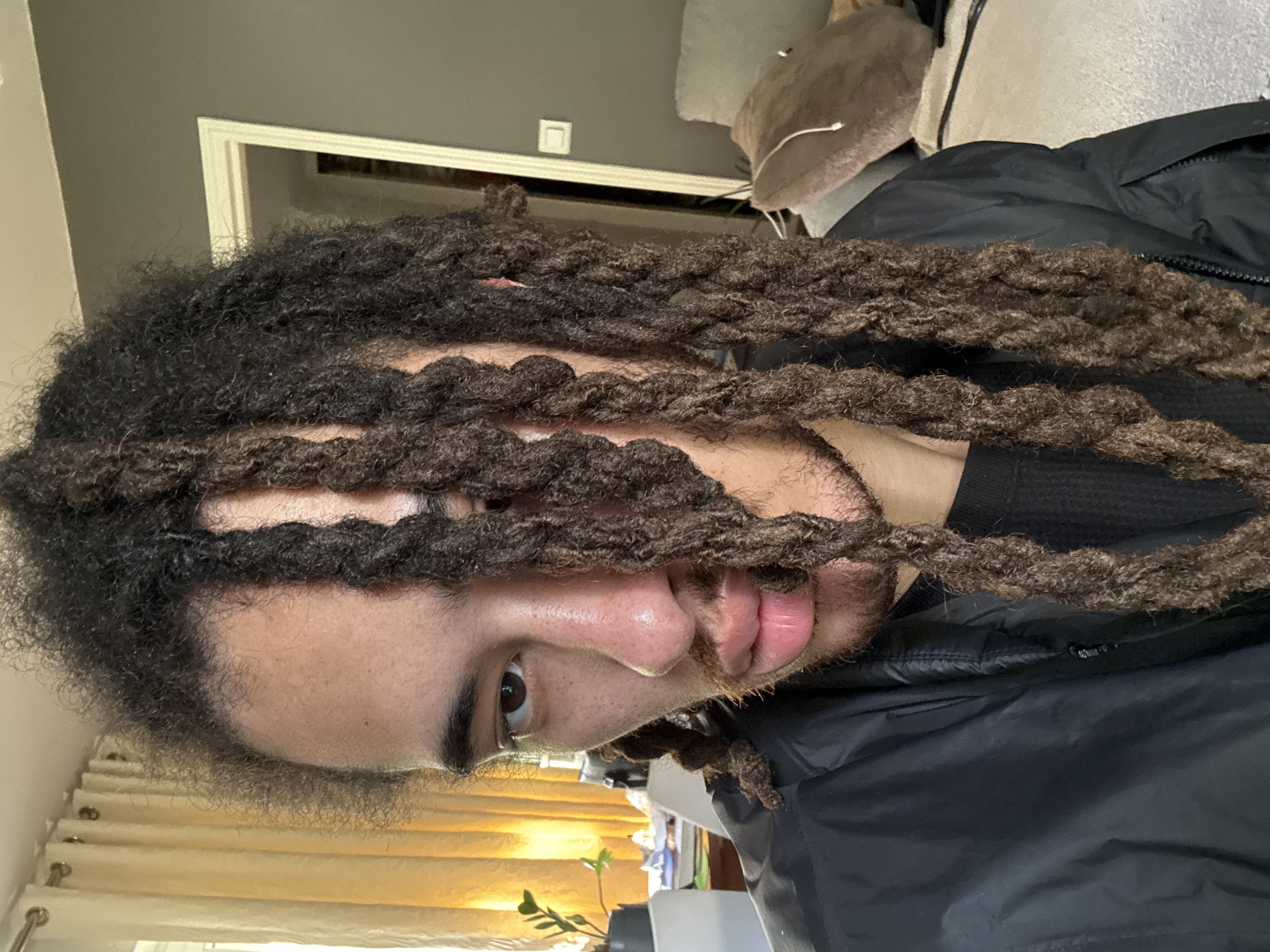 A person with dark hair styled in dreadlocks is positioned close to the camera. The dreadlocks partially obscure their face, especially the eyes and forehead. The person has brown eyes, a light brown complexion, a mustache, and a beard. They wear a black jacket and a black shirt. A window with yellow curtains is visible in the background, along with a houseplant, a door frame, and a light switch. The location is unspecified, and there is no discernible sign indicating the city of Les Ulis, France. There are no detectable actions or interactions in the scene.