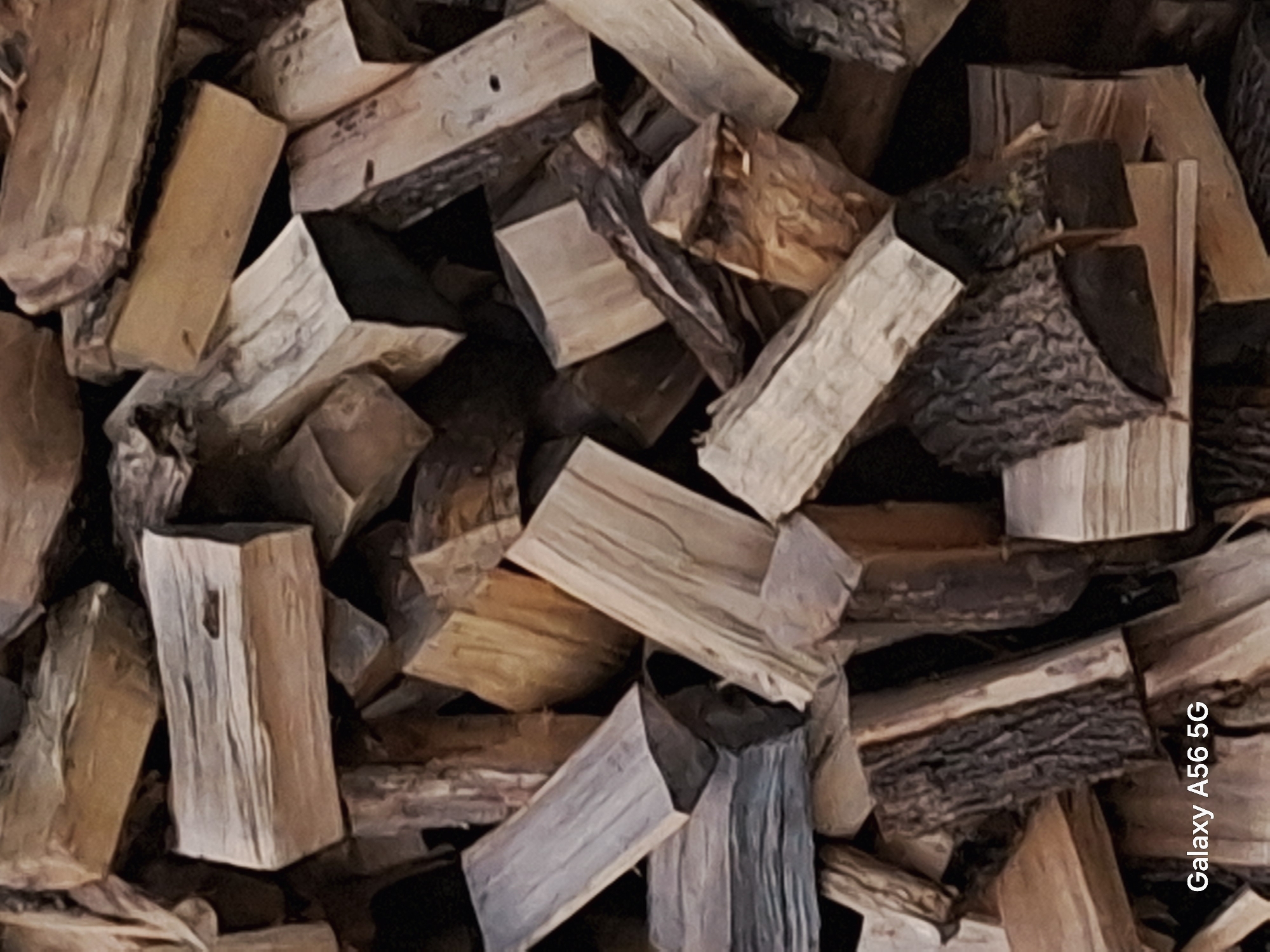 The image displays a close-up, overhead view of a large pile of chopped firewood. The wood is stacked haphazardly, with pieces of varying lengths and thicknesses. The texture of the wood is rough and varied, with some pieces showing smooth, cut ends and others displaying the rough bark of the trees. The colors range from light tan and beige to darker browns and grays. The lighting appears natural, casting shadows that highlight the three-dimensional nature of the woodpile. The bottom left corner of the image contains the text "Galaxy A56 5G". There are no people or animals visible in the image. The scene is consistent with a storage area for firewood, likely in a rural or semi-rural setting. This image does not contain information about the city of Saliai, Lithuania.