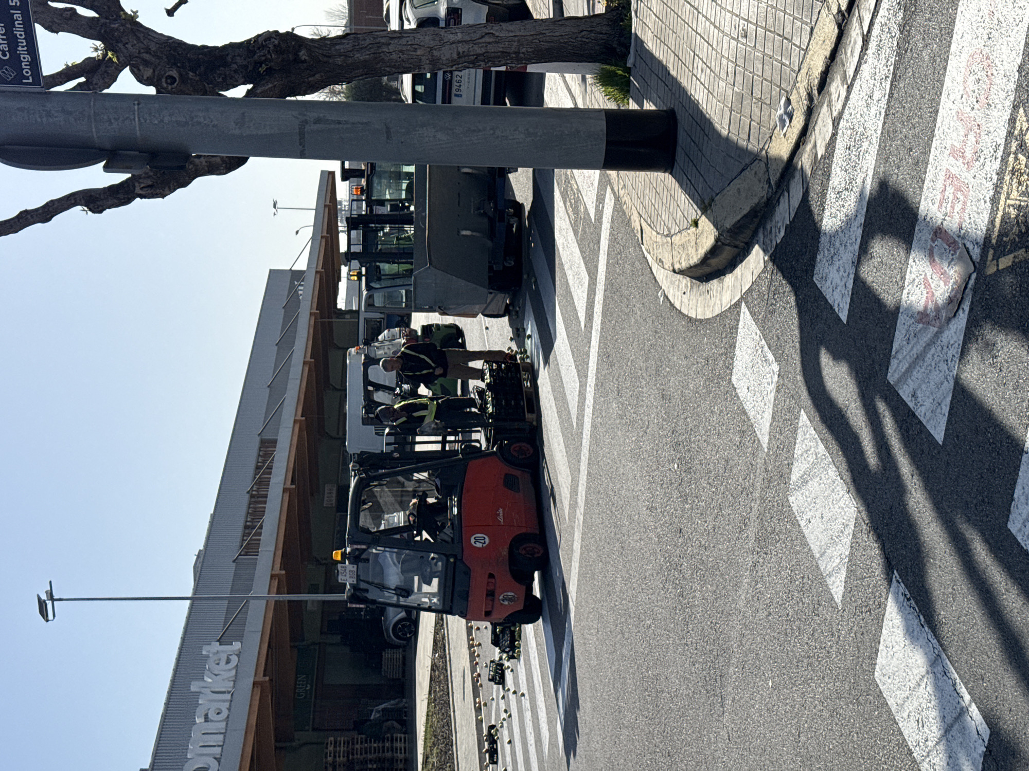 The image depicts a street scene in Barcelona, Spain. A red Linde forklift, its license plate partially visible as "E 6549 BGB," is positioned on a zebra crossing in front of a supermarket, likely Green Market, as indicated by a sign. The street name "Carrer Longitudinal 5" is visible on a street sign attached to a pole. Debris, possibly produce, is scattered on the crossing. Several people are visible, including the forklift operator and other workers on or near the forklift. In the background, other vehicles and commercial buildings are present. A tree partially obstructs the upper left area of the frame, casting a shadow on the zebra crossing. The day appears bright, with clear skies.