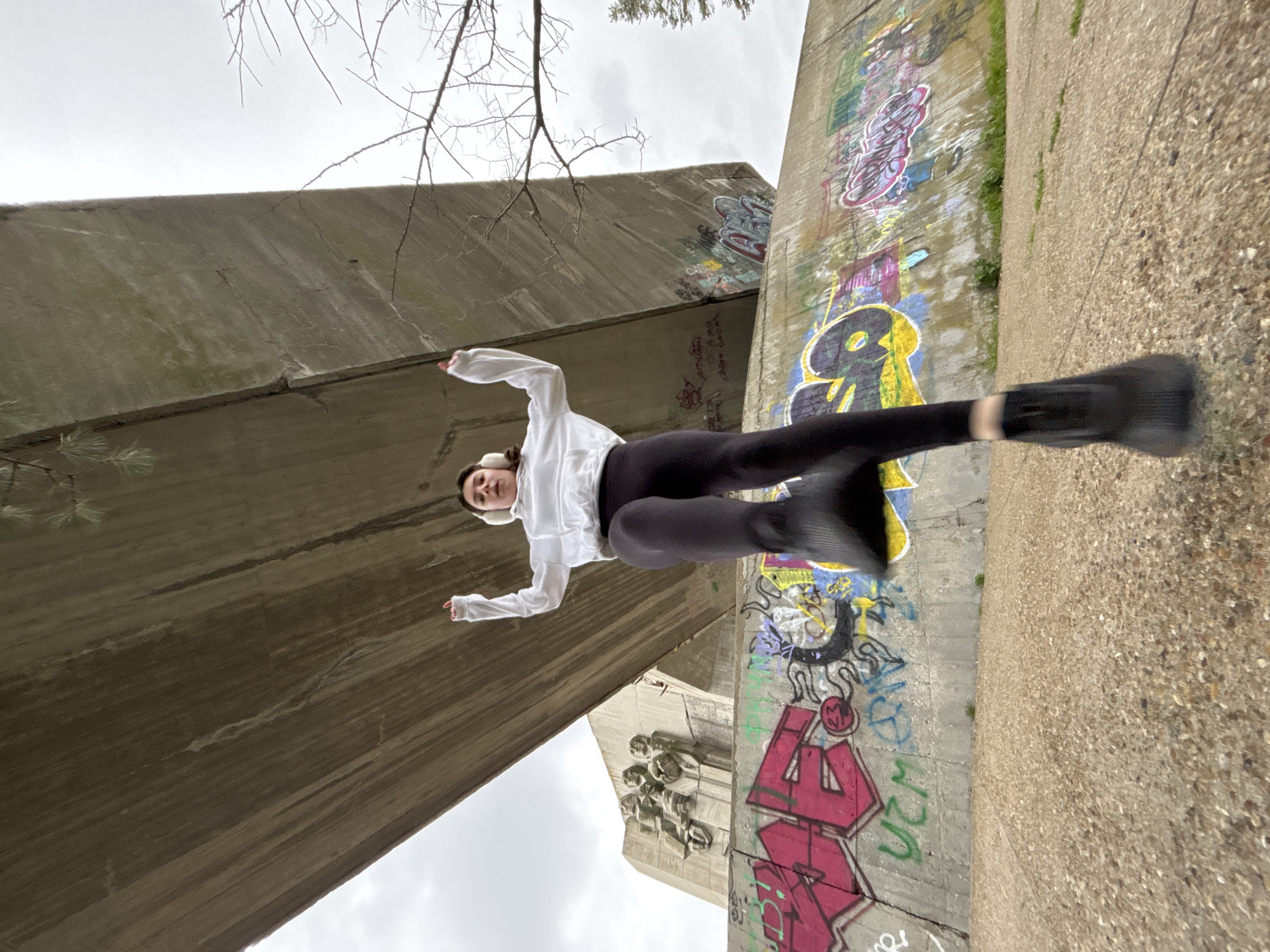 A low-angle shot depicts a female in mid-air, appearing to jump. She wears a white long-sleeved shirt, black leggings, and black sneakers. She has white earmuffs on her head. The background includes concrete structures covered in graffiti. A cloudy sky is visible in the upper left corner. A tree branch is visible in the upper center. A concrete relief carving is visible below the female. The city and specific location are unknown.