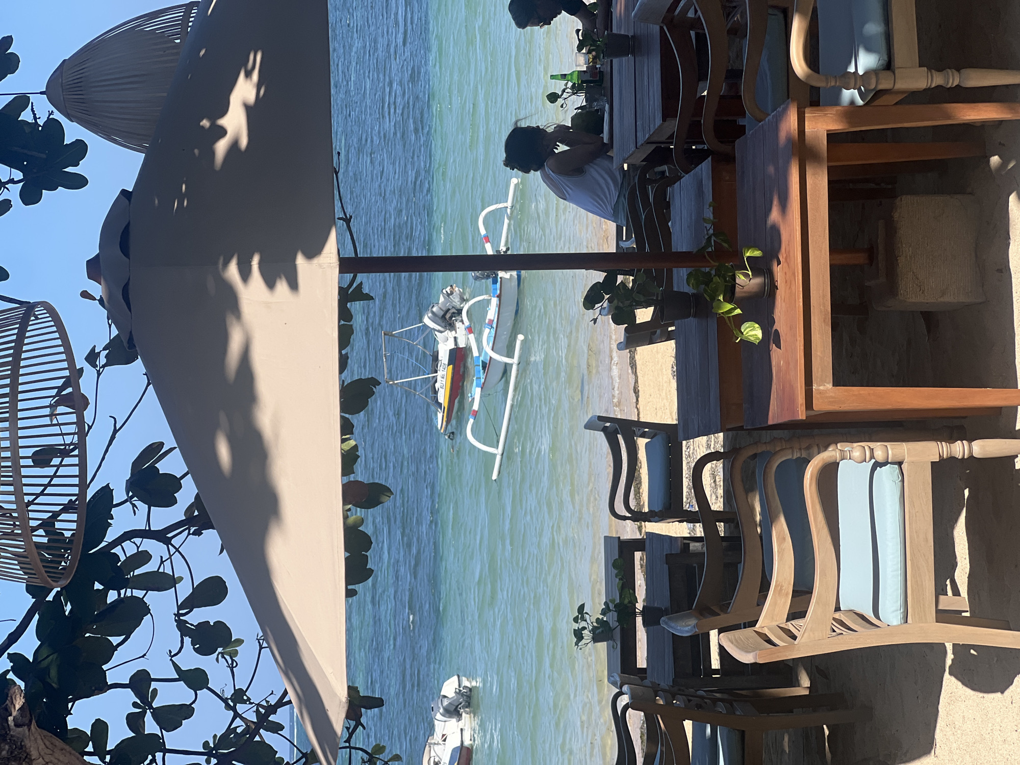 An outdoor dining area is set up on a sandy beach overlooking a turquoise ocean. Several wooden tables and chairs with light blue cushions are arranged under a large, light-colored umbrella. The tables are set with potted plants. In the background, two boats are anchored in the water, one a small white motorboat and the other a traditional outrigger canoe. Two individuals are seated at a table in the mid-ground, facing away from the camera. One person is wearing a white tank top and the other is wearing a dark shirt. The sky is clear and blue, and the sun casts shadows from the umbrella and overhead foliage onto the scene. The location is identified as Denpasar, Indonesia.