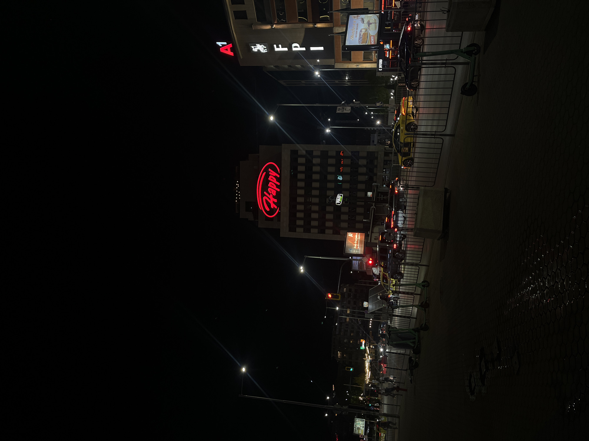 At night, a cityscape in Sofia, Bulgaria is illuminated by streetlights and building signs. Prominently featured are the "Happy" restaurant sign in red, and the "A1 EPI" building sign. Cars are seen in traffic on the street. The sidewalk features parked scooters.