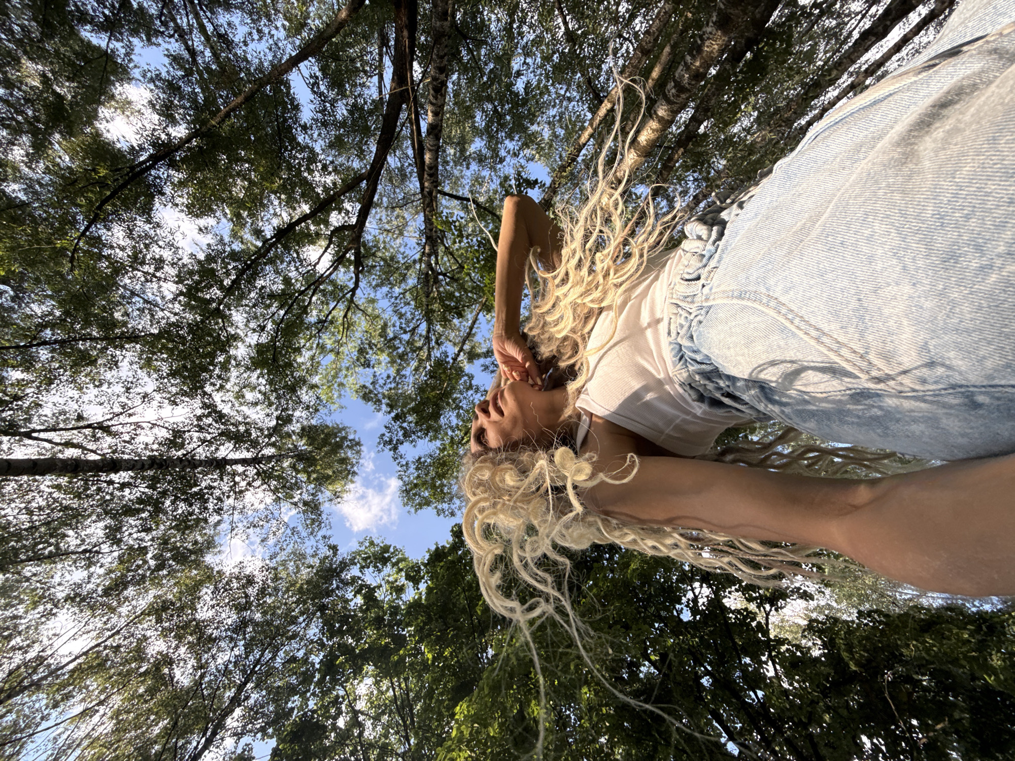 A low-angle shot captures a blonde woman with long, curly dreadlocks against a backdrop of a blue sky and lush green trees. Her white tank top and light-wash denim skirt create a casual summer outfit. She looks upwards, her head tilted back and hand resting on her neck, her hair flowing around her. The sunlight filters through the leaves, casting dappled shadows.