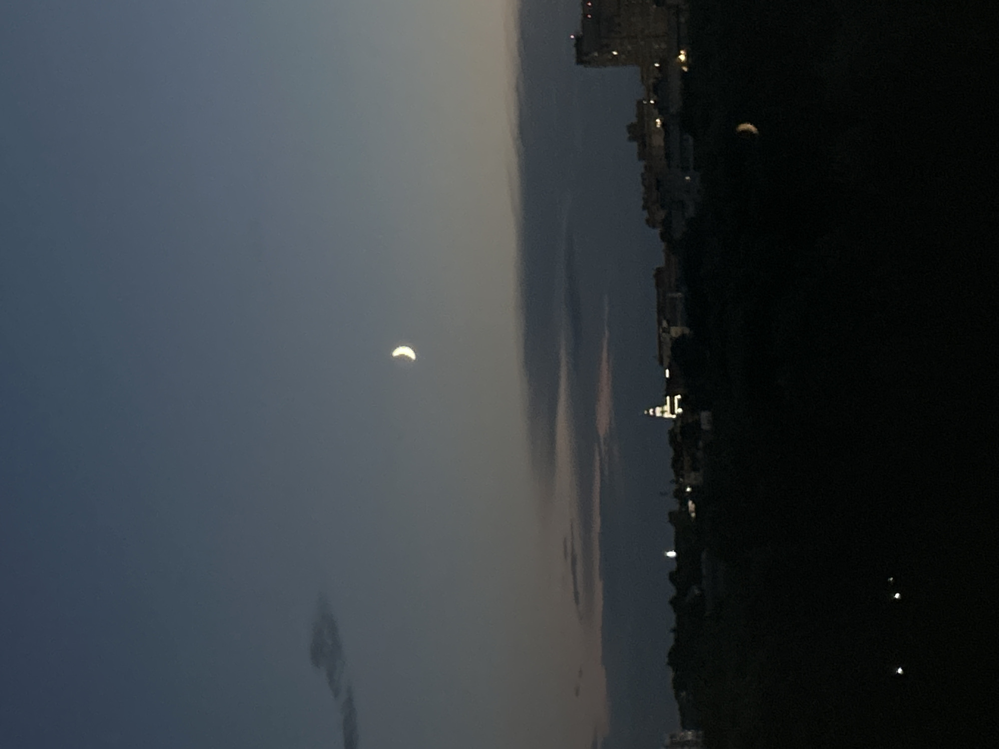 The image captures a crescent moon hanging in a dusky, deep blue sky over a city skyline. The sky is partly cloudy with a few wispy clouds visible. Below the sky, a dark silhouette of trees and hills separates the sky from the cityscape. The city itself is illuminated with scattered lights from buildings. A prominent building with lights on is visible on the right side of the image. A tower or spire with illuminated windows is also visible within the city. The foreground is very dark, obscuring any details of the ground. The overall scene is a twilight view of a city under a moonlit sky.