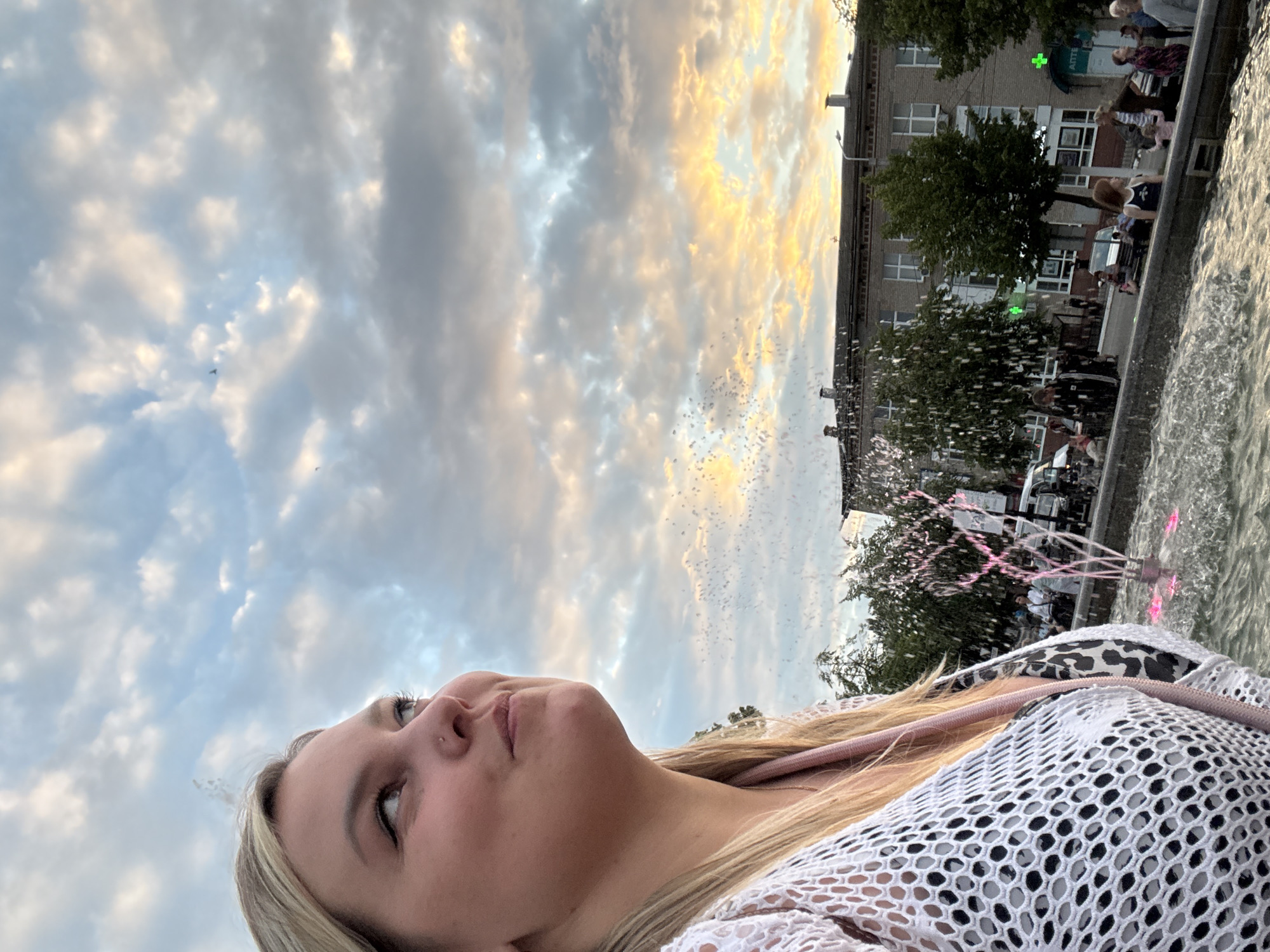 This is a medium shot, slightly angled, outdoor photo of a young woman in the foreground. She has blonde hair, light skin, and is looking upwards and to her left with a slight smile. She is wearing a white, open-knit top over a black and white patterned garment. A pink strap is visible across her chest. In the background, there is a fountain with pink lights, and water spraying upwards. Beyond the fountain, there is a street with buildings, trees, and people. The sky above is filled with dramatic, cloudy textures and a sunset glow in the distance. The location is the fountain area on Promyshlennaya Street in Chaikovsky, Russia.