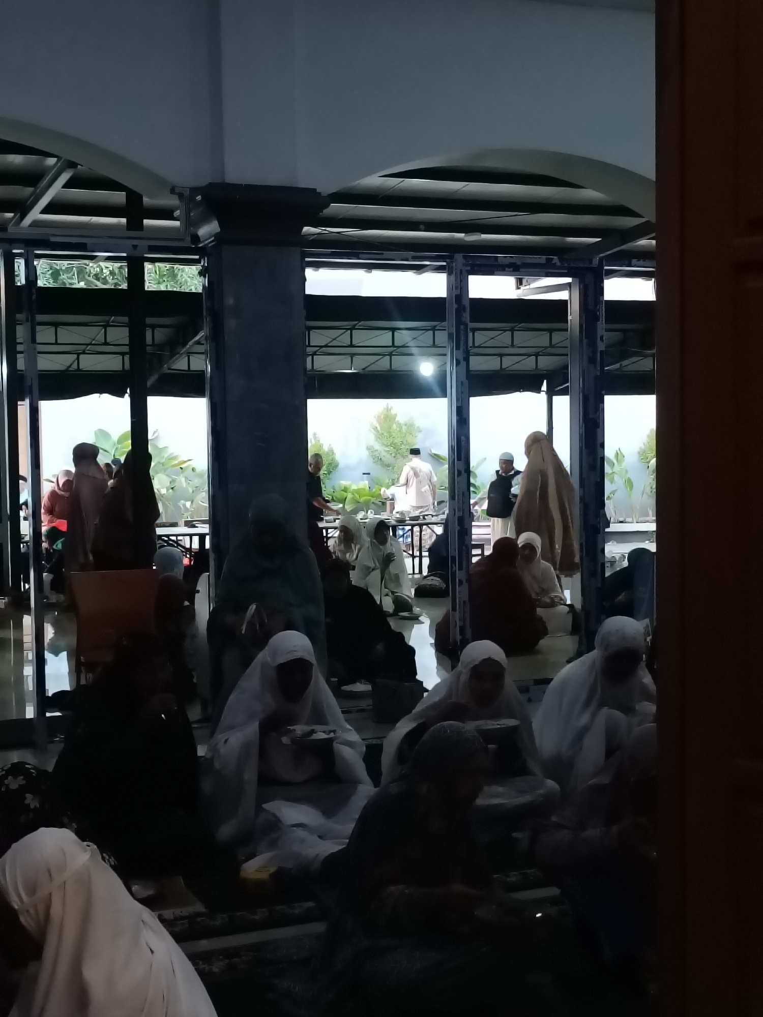 The image depicts a group of people, predominantly women wearing hijabs and traditional Islamic attire, gathered indoors. They are seated on the floor, some appearing to be engaged in prayer or a religious gathering. The environment suggests a place of worship or a community hall, possibly a mosque. The lighting is dim, indicating it might be evening or a poorly lit indoor space. The architectural elements visible, such as columns and a covered outdoor area visible through glass panels, hint at a modern construction style within a tropical or subtropical climate, as suggested by the lush greenery outside. The presence of what appear to be serving trays or plates with food in front of some individuals indicates a communal meal or event. The overall scene conveys a sense of religious observance and community.