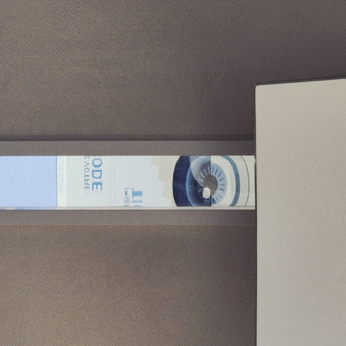 The image displays a narrow, vertical, reflective strip, likely a mirror or highly polished surface, situated centrally within the frame. This strip is positioned against a textured, brownish-grey background, which appears to be a wall or partition. Reflected within the strip, and therefore appearing mirrored and inverted, are elements of a display or sign. Visible French text includes "VOTRE" and "CODE", rendered in blue on a white background, above characters that appear to be "IT". Below this text, a stylized graphic of a human eye, depicted in shades of blue and white with a dark central pupil, is also reflected. The lower portion of the image reveals a light grey, flat horizontal surface, consistent with a desk or counter, extending across the foreground. The scene is an interior environment, indicative of a public or semi-public facility featuring self-service terminals. Given the reflected French text "VOTRE CODE" and the characteristic eye graphic, this scene is identified as an interactive terminal within a Pôle Emploi agency located in Strasbourg, France. No individuals or active interactions are present in the image.