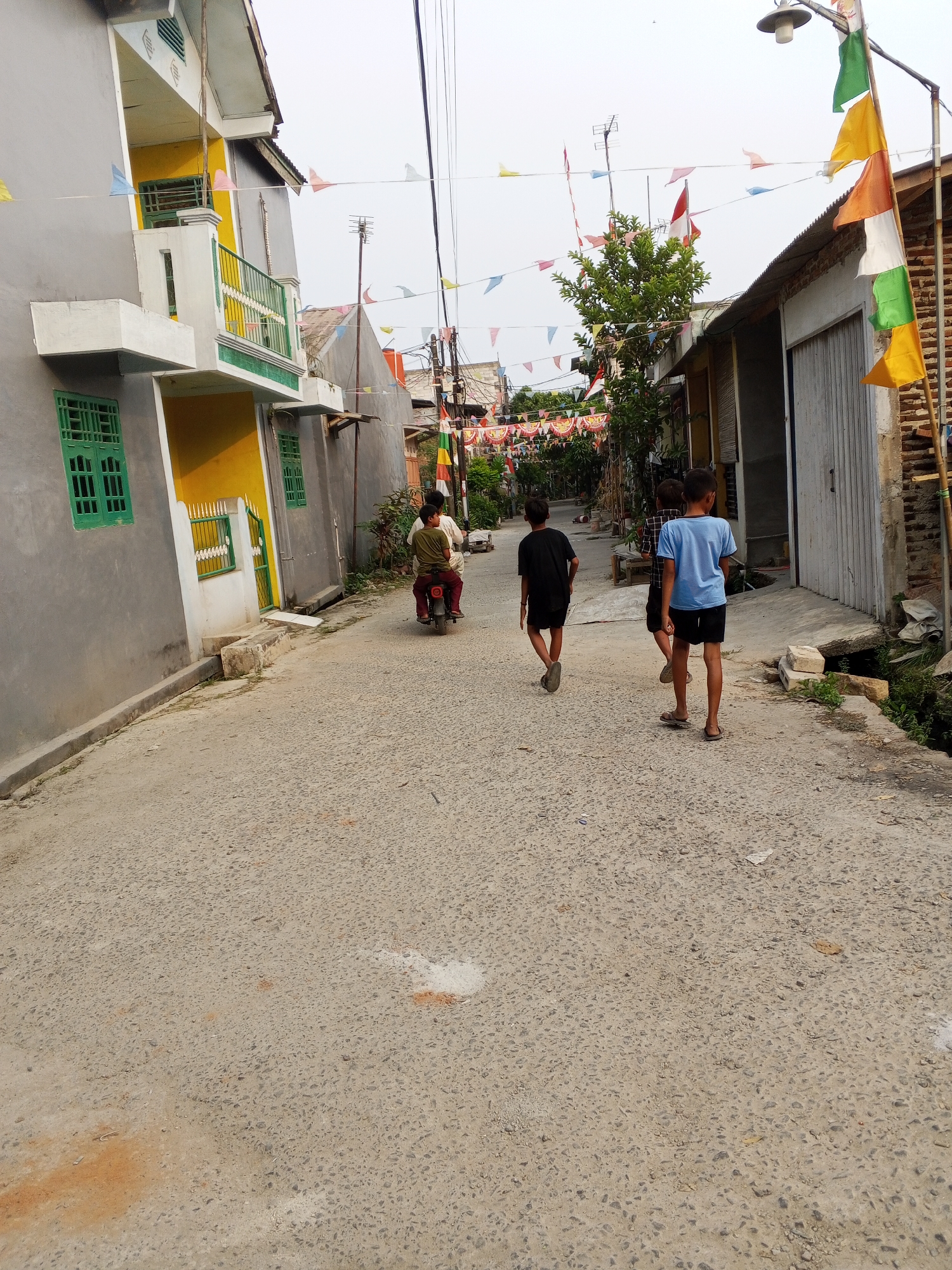 The image depicts a narrow, paved street in Ciruas, Indonesia, lined with residential buildings and decorated with numerous colorful triangular flags. On the left side of the street, a two-story house with grey and yellow walls and green window frames is visible. The house features a balcony with a green railing on the upper floor. The right side of the street shows a building with a corrugated metal roof and a large sliding door. In the middle of the street, a person is riding a red motorcycle, and three young boys are walking away from the camera. The boys are wearing casual clothing: one in a black t-shirt and shorts, another in a blue t-shirt and shorts, and a third in a plaid shirt and shorts. The sky is overcast. The scene appears to be set during daylight hours, with the flags suggesting a celebratory occasion.