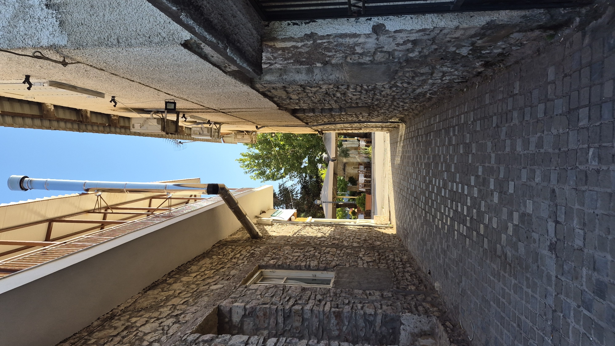 A narrow cobblestone alleyway is depicted, flanked by stone buildings on both sides. The building on the left features a balcony with a wooden railing and a prominent stainless steel exhaust pipe running vertically along its exterior. The building on the right has a textured, light-colored facade with visible air conditioning units and wiring. At the end of the alley, an outdoor seating area with tables and umbrellas is visible, suggesting a cafe or restaurant. Lush green trees are present beyond the seating area. The scene is illuminated by natural daylight, casting shadows that emphasize the textures of the stone walls and the cobblestone pavement.