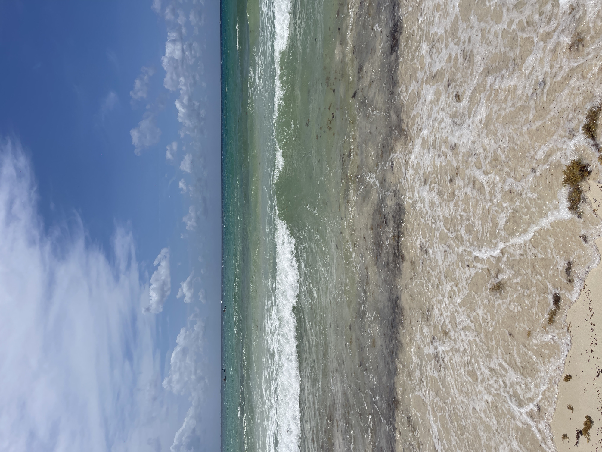 The photograph captures a beach scene in Playa del Carmen, Mexico. The view extends from the sky to the shoreline. The left third of the image shows a blue sky dotted with fluffy white clouds. The middle third is of the ocean with small white waves breaking near the shore. The right third of the image is of a sandy beach with foamy water washing over it. Bits of seaweed are scattered along the beach and in the shallow water. The picture is taken from an elevated perspective.
