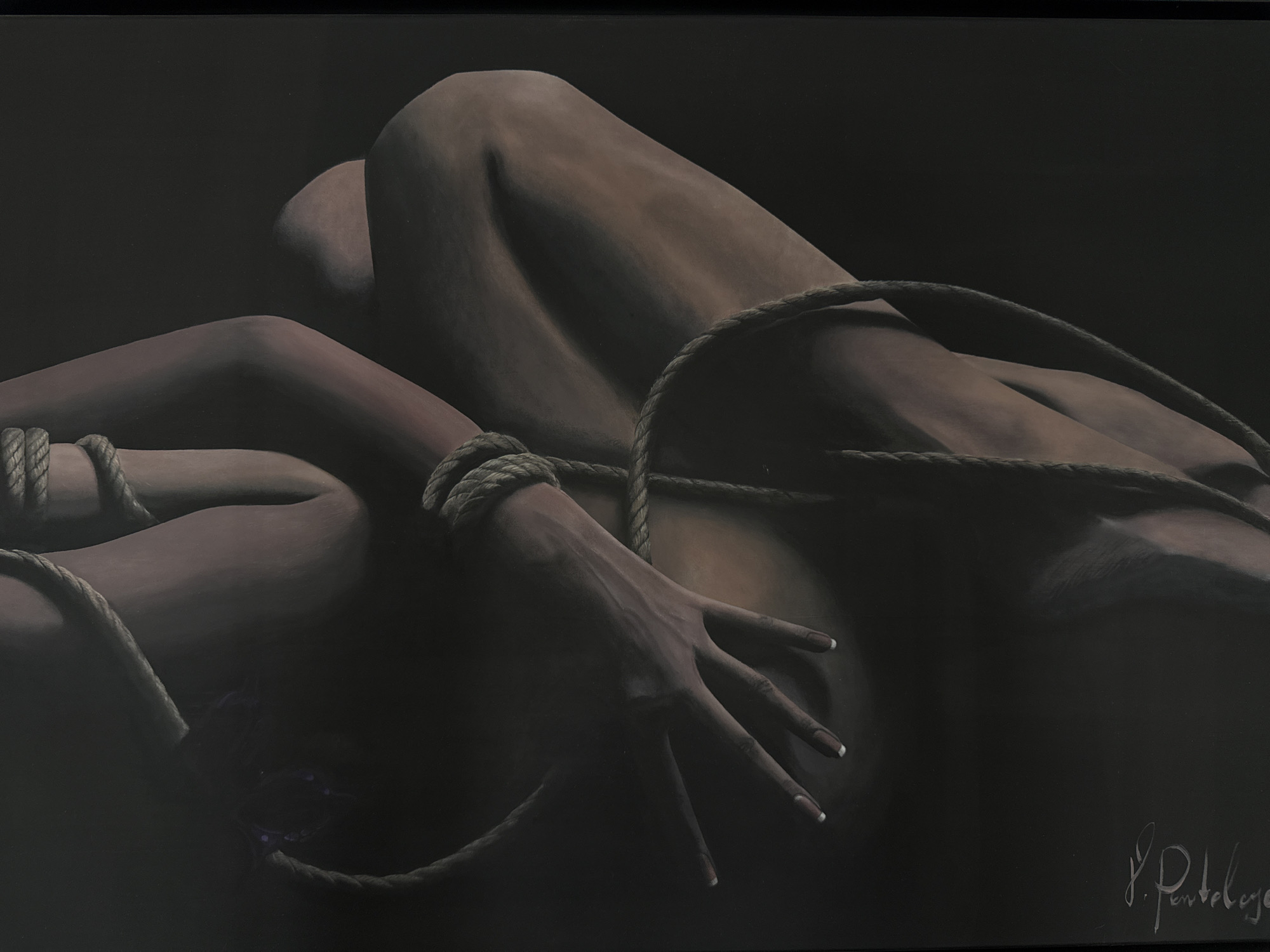 The image is a close-up, low-angle shot of a person's body, bound with thick rope. The person is lying down, with their legs bent and their arms crossed across their torso. The rope is tightly wrapped around their wrists and forearms, and also appears to be securing their legs. The lighting is dim, casting long shadows and highlighting the contours of the body. The skin tone is a warm brown, and the rope is a natural fiber, likely jute or hemp. There is a signature in the lower right corner of the image, which appears to read "P. Panteloye." The background is a dark, nondescript surface. There is no specific indication of location in Mesogi, Cyprus.