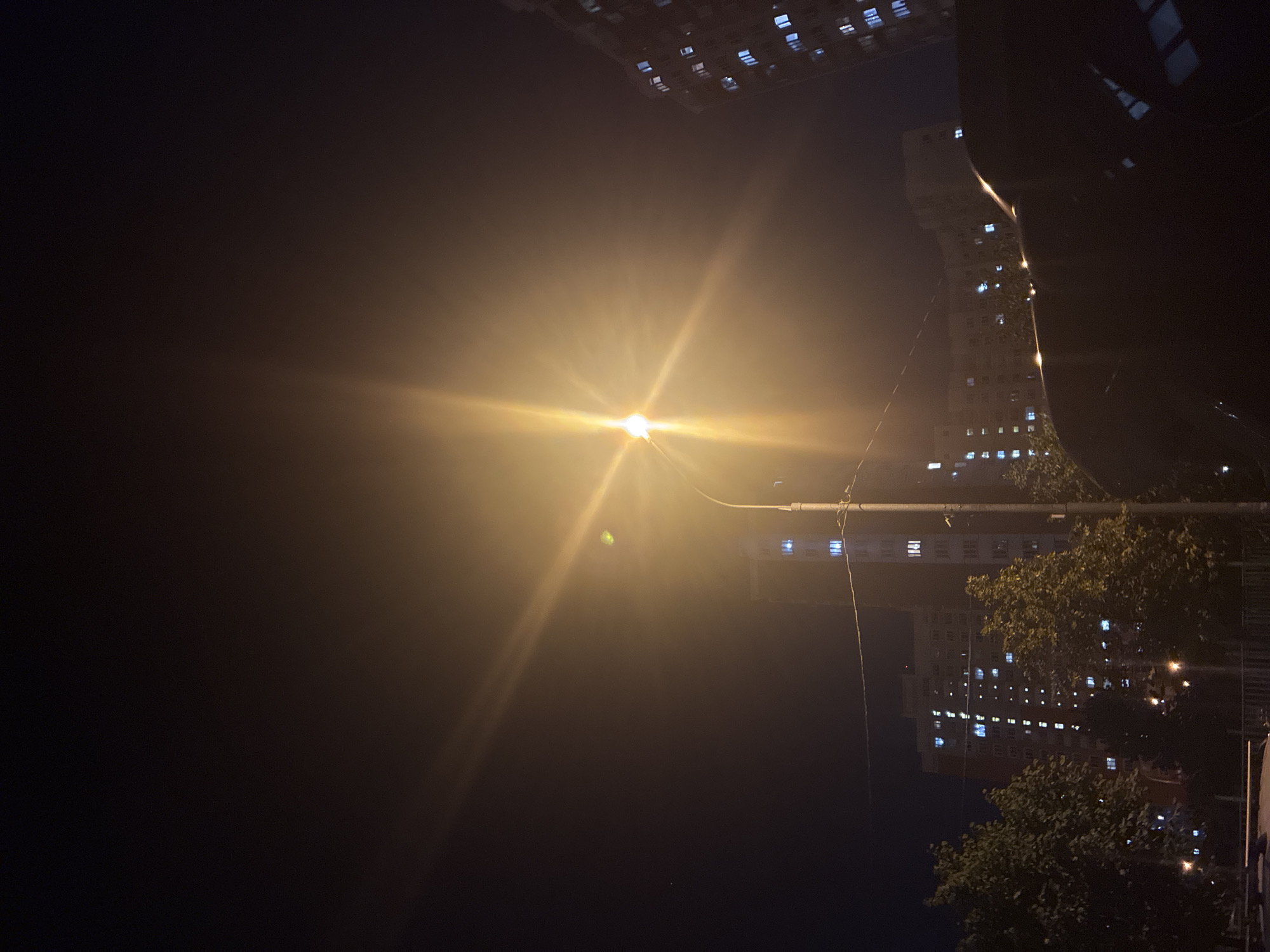 The image depicts a nighttime street scene in Drozhzhino, Russia. A bright street lamp dominates the upper center of the frame, casting a strong glare and lens flare across the dark sky. Several multi-story apartment buildings are visible in the background, their windows illuminated with blue and white light, suggesting occupancy. Trees with dark foliage are scattered in the mid-ground, obscuring parts of the buildings. In the foreground, the dark silhouette of a vehicle, possibly a car, occupies the lower portion of the image. The overall atmosphere is urban and dimly lit, illuminated primarily by artificial light sources. No people are directly visible.