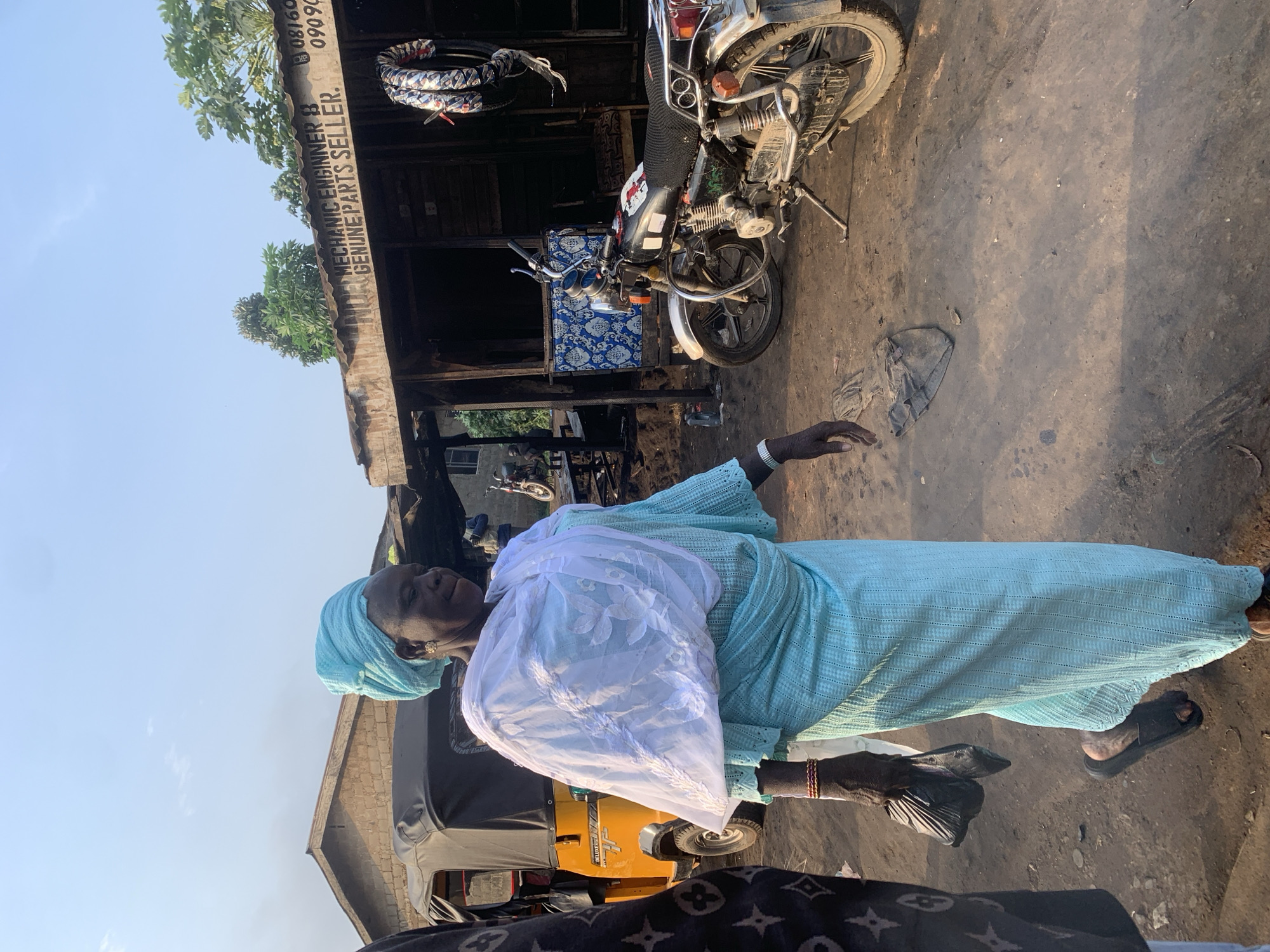 In the city of Shagamu, Nigeria, a woman is walking past a mechanic shop. She is wearing a light blue traditional dress and a matching headscarf, with a white translucent shawl draped over her shoulders. She carries a black plastic bag in her left hand. To her left, a yellow auto-rickshaw is parked. The mechanic shop, constructed with corrugated metal roofing and wooden planks, has a sign above its entrance that reads "MECHANIC ENGINNER B GENUINE PARTS SELLER" and phone numbers. A motorcycle is parked in front of the shop, and several tires are displayed on the wall. The ground is unpaved and dusty, with tire tracks visible. The overall scene depicts a bustling street in Shagamu.
