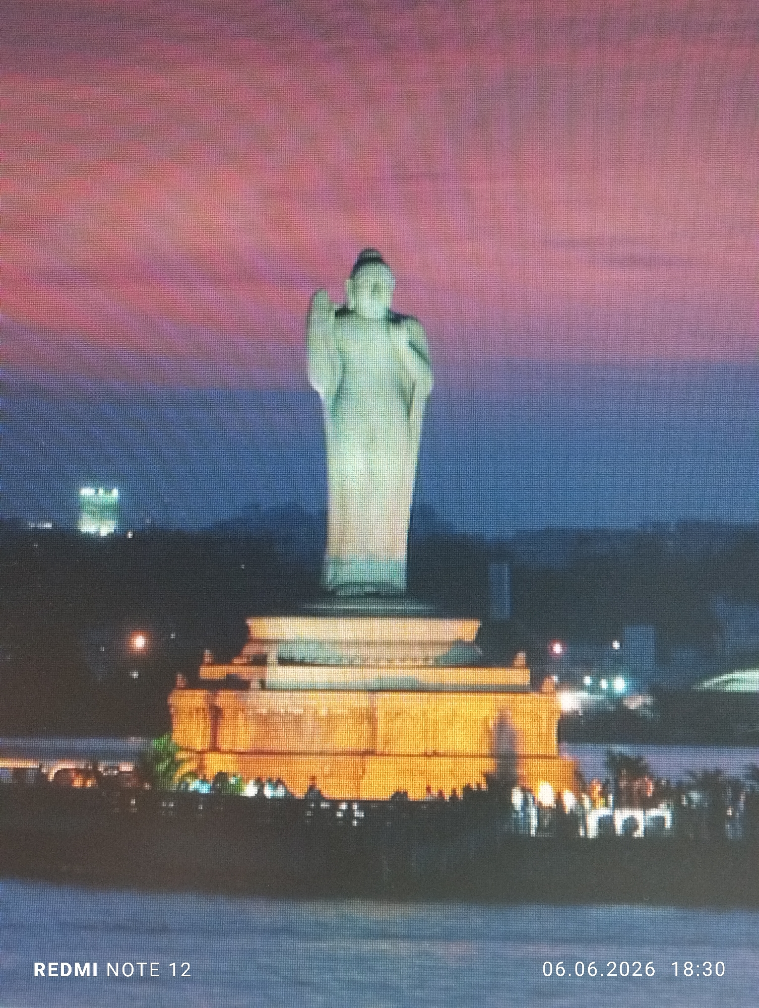 The image presents a large, light-colored standing Buddha statue centrally positioned within a body of water, likely a lake, captured during twilight. The statue's multi-tiered base is prominently illuminated with warm, golden light, revealing numerous silhouetted individuals gathered on the surrounding platform. The sky above transitions dramatically from deep blue at the horizon to vibrant pink and purple hues. In the distant background, city lights and faint building silhouettes are discernible against the dark landscape. The entire image displays a distinct grid pattern, suggesting it may be a photograph of a digital screen or a highly pixelated digital file. Overlaid white text in the bottom left corner reads "REDMI NOTE 12," while the bottom right corner displays "06.06.2026 18:30." While the request specifies the location as Burgas, Bulgaria, the monument depicted is unequivocally identified as the Buddha statue located in Hussain Sagar Lake, Hyderabad, India, and is not a known landmark in Burgas, Bulgaria.