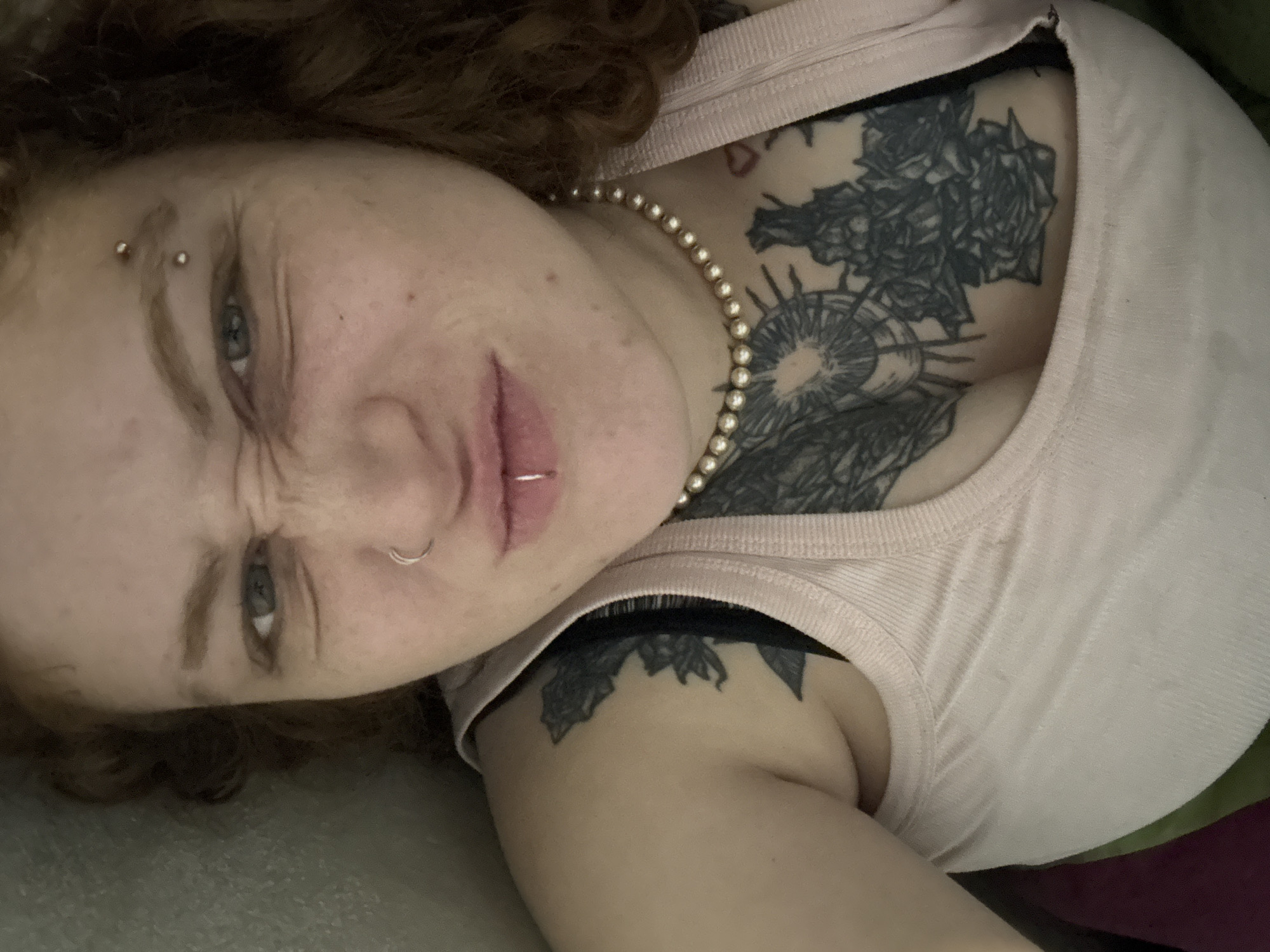 A close-up, angled portrait of a person with fair skin and reddish-brown curly hair, possibly female, is presented. The person is wearing a light pink tank top with what appears to be black lingerie visible at the neckline. A pearl necklace adorns their neck. Several piercings are evident: one in the upper right eyebrow, one in the left nostril, and one through the lower lip. Tattoos are visible on the upper chest and shoulder. The background is out of focus, suggesting the person is lying down on a soft surface. The scene is NOT demonstrably identifiable as being located in Buenos Aires, Argentina.