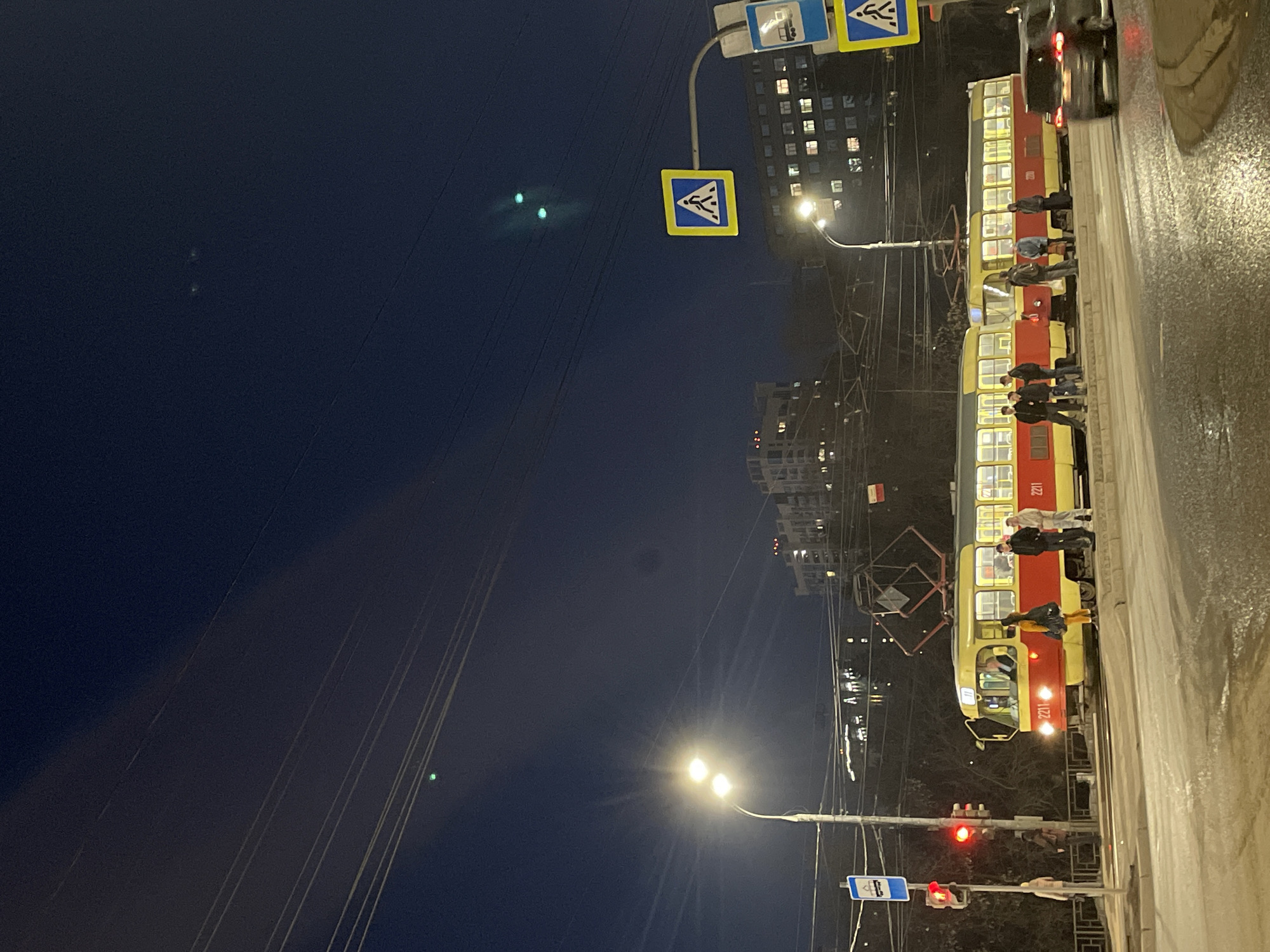 The image shows a street scene at dusk or night in Izhevsk, Russia. A red and yellow tram is stopped on the right side of the frame, with passengers standing at its open doors. Some people are on the sidewalk next to the tram.
The street is wet, reflecting the lights from street lamps and the tram. On the left, the sky is a deep blue, with power lines visible against it. In the upper portion of the image, there are road signs, and beyond them are buildings with lit windows. The streetlights are emitting a bright glow. A traffic signal is seen near the bottom center of the picture, displaying red.