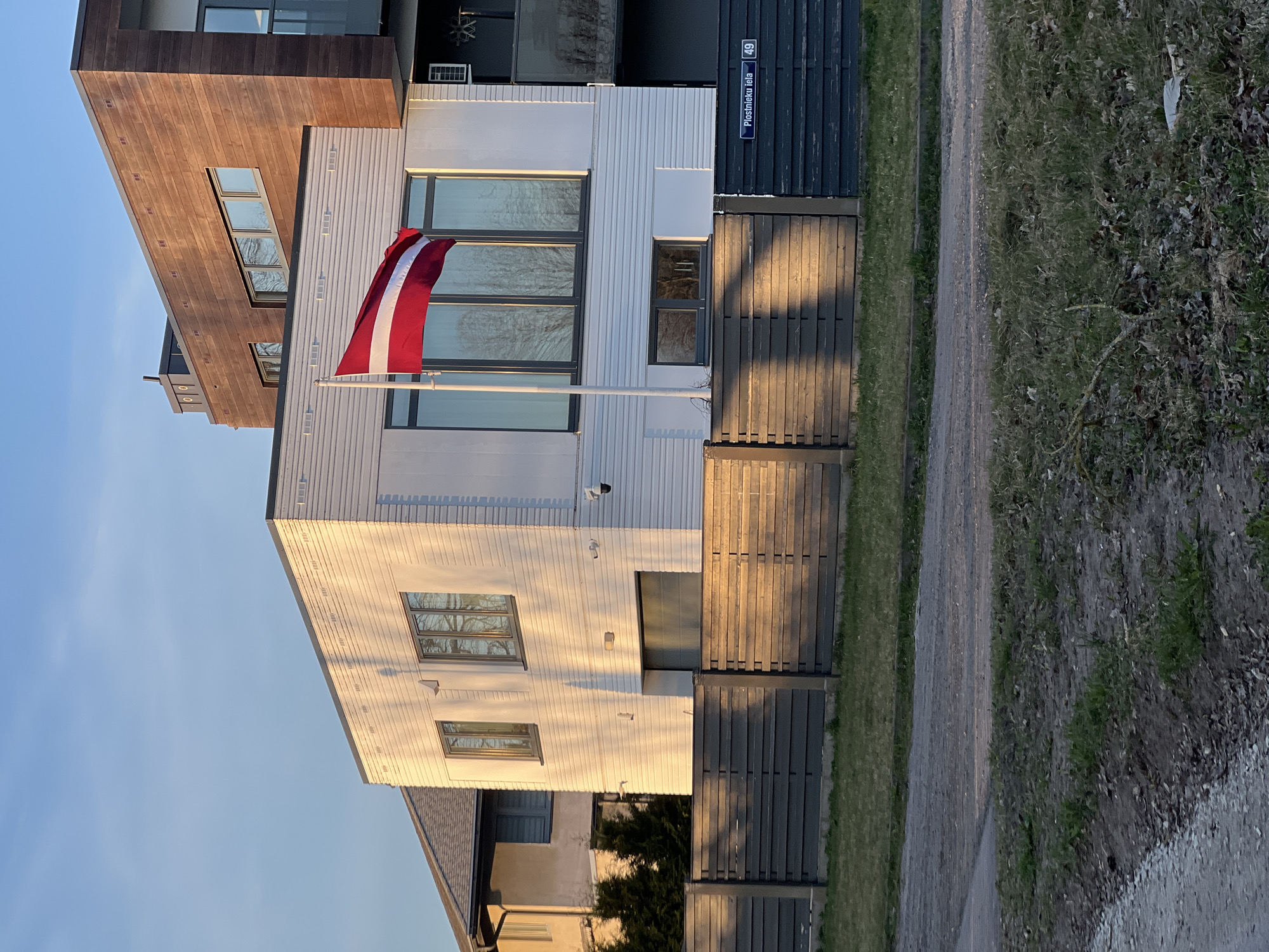 The image shows a modern building in Riga, Latvia, identified by a street sign reading "Plostnieku iela 49." A Latvian flag is prominently displayed on a flagpole attached to the building. The building has a mix of white and dark wood cladding, with large windows reflecting the surrounding environment. A dark wooden fence runs along the property line, adjacent to a gravel path and a strip of grass. The sky is light blue, suggesting it is either early morning or late afternoon. The image is taken from an angle that includes a section of the path and vegetation on the right side. A partial view of another building is visible in the lower left corner.