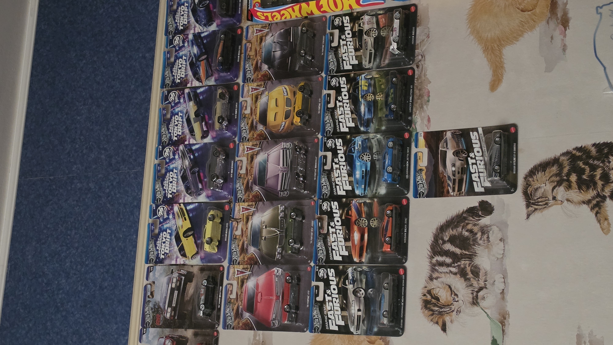 The image shows a collection of Hot Wheels "Fast & Furious" and "Tokyo Drift" die-cast cars in their original packaging. The cars are arranged in rows and columns on a surface featuring a printed design of cats. The image does not appear to be taken in Varna, Bulgaria, nor does it show any identifiable location. There are no people visible in the image. The focus is solely on the collection of Hot Wheels cars.