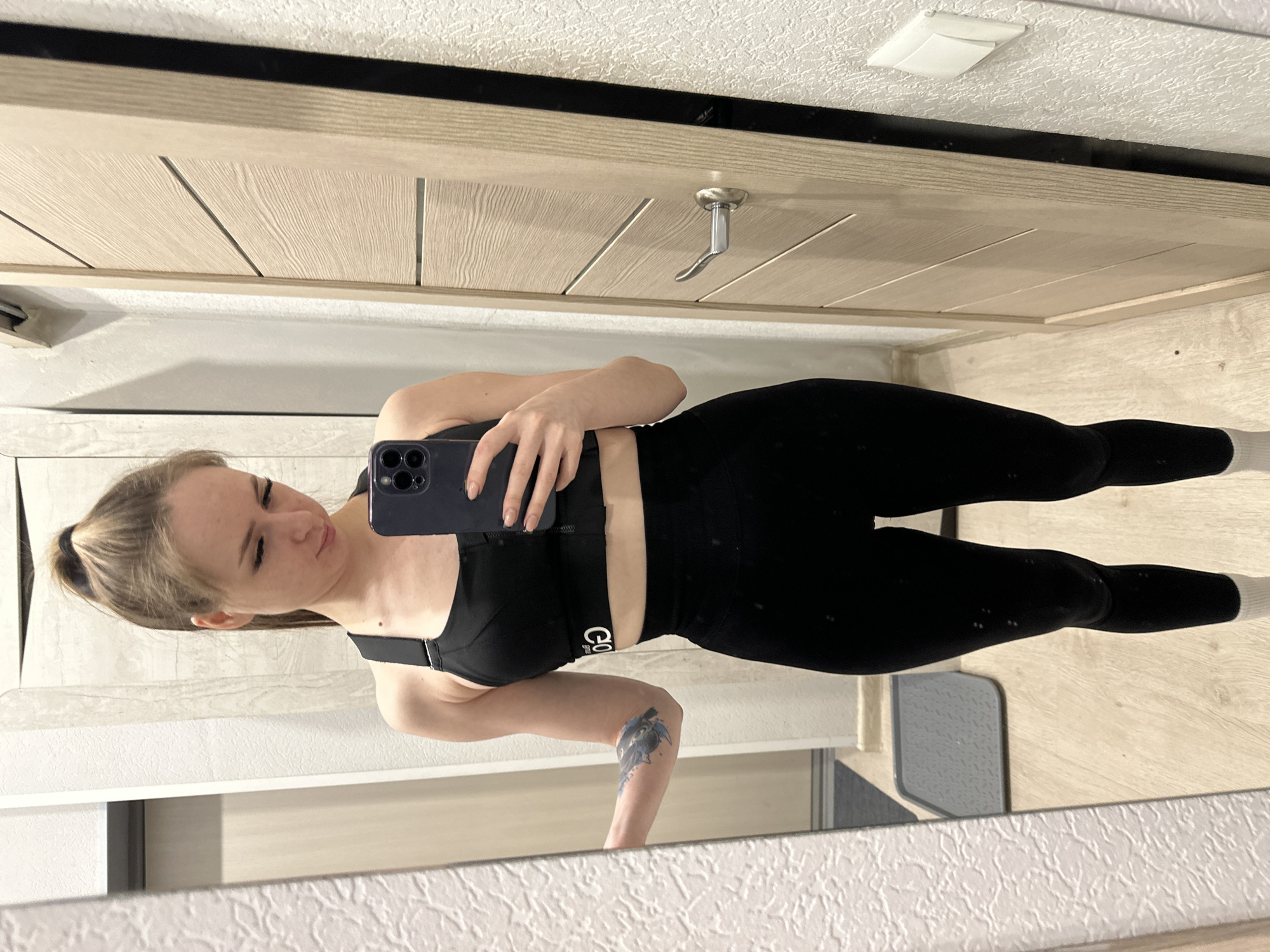 A woman stands in front of a mirror, holding a phone to take a selfie. She is wearing a black sports bra and black leggings. The room appears to be a hallway with a closed door and white walls. The location is Unknown, Unknown.