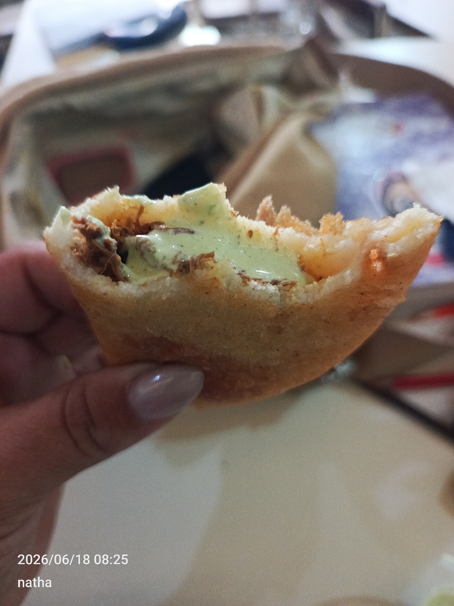 A close-up shot shows a person's hand holding a half-eaten, deep-fried pastry filled with what appears to be seasoned meat and a creamy green sauce. The pastry is golden brown and slightly irregular in shape, with a crispy texture visible along its edges. The filling is a mix of shredded brown meat and a dollop of light green sauce. The person's fingernail, which is painted in a neutral tone, is visible on the left side of the frame, indicating a focus on the food item. In the blurred background, elements of a casual setting can be seen, including what looks like a bag or container and other indistinct objects. The lighting suggests an indoor environment, possibly a restaurant or home, during the day. A timestamp in the lower left corner reads "2026/06/18 08:25" and the word "natha" is also visible, likely identifying the creator or subject of the image. The overall impression is of someone enjoying a snack, captured in a moment of casual dining.