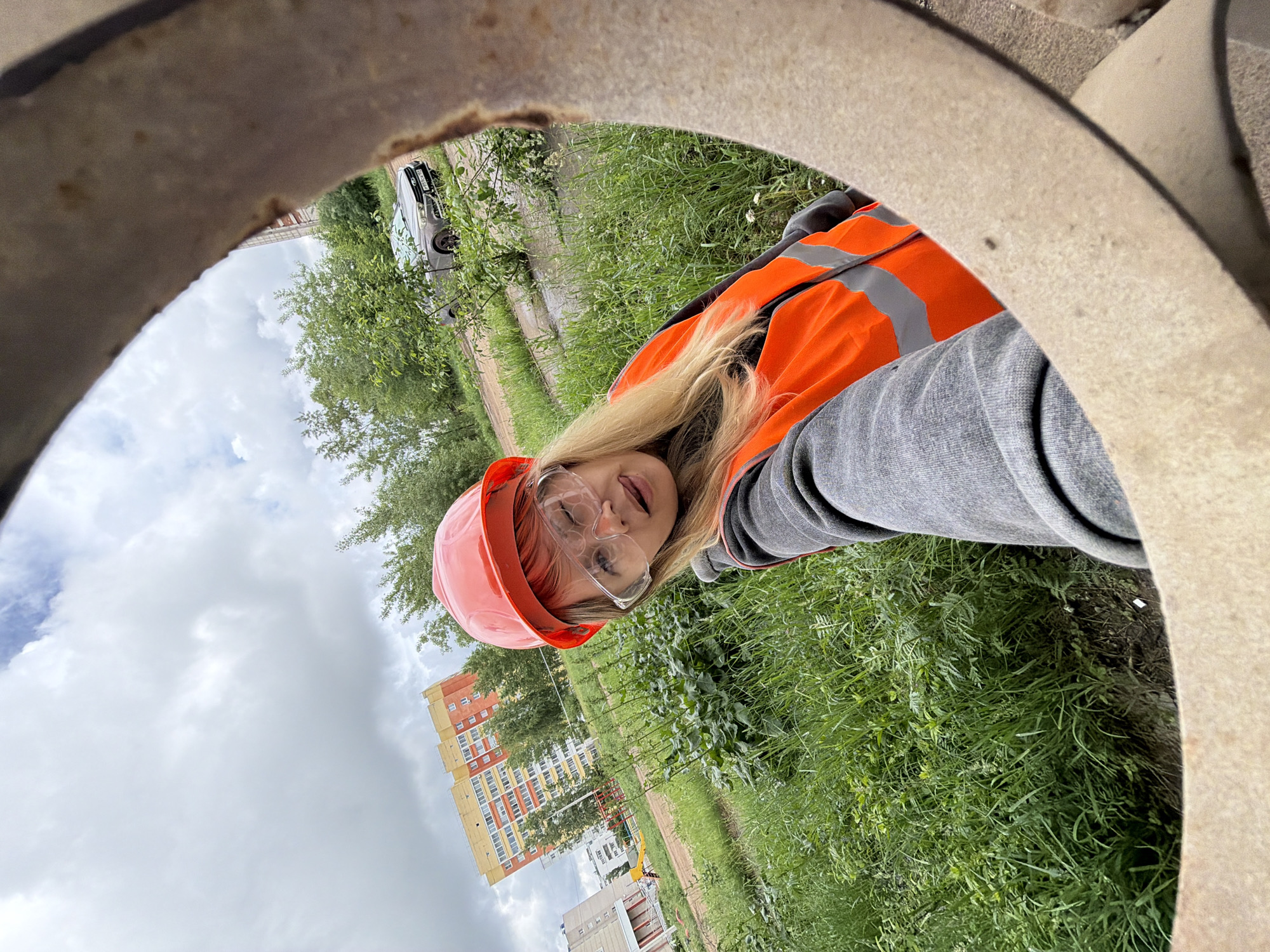 A young woman with blonde hair is captured in a selfie-style photograph, peering out from inside a large, circular concrete or stone opening. She wears an orange hard hat, clear safety glasses, and an orange high-visibility vest over a grey long-sleeved top. Her expression is cheerful, with a slight smile and her tongue briefly visible as she looks directly at the camera.
The background, framed by the rough edges of the opening, reveals an outdoor setting. Lush green grass and trees are prominent, suggesting a temperate season. A dark-colored sedan is parked on a dirt path in the midground. Further in the distance, a multi-story residential building, characterized by yellow and orange facades, stands beneath an overcast sky with scattered clouds. Other smaller buildings and structures are also visible. The scene suggests a location like a construction site, an industrial area, or a public green space within a city, consistent with a setting in Chaikovsky, Russia. The lighting indicates it is daytime, with diffused light from the cloudy conditions. No discernible text is visible anywhere in the image.