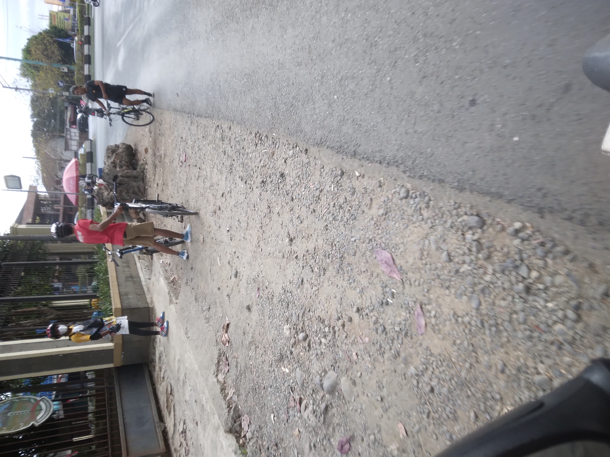 Three people are visible near a paved road with a gravel shoulder in Desa Pamijen, Kecamatan Sokaraja, Indonesia. The person on the left is wearing a white shirt with yellow accents, a white skirt, black leggings, a red and black helmet, and a white face mask. This individual is standing beside a bicycle with a blue frame and is facing to the right. To the right of this person is another individual wearing a red t-shirt and tan shorts, also with a helmet, standing with a bicycle. Further to the right, a third person is wearing a black t-shirt and black shorts, holding a bicycle and facing left. A large grey rock formation is situated behind the two people closer to the left. The environment appears to be outdoors with some vegetation visible in the background, and buildings line the far side of the road. The road is wet, suggesting recent rain.