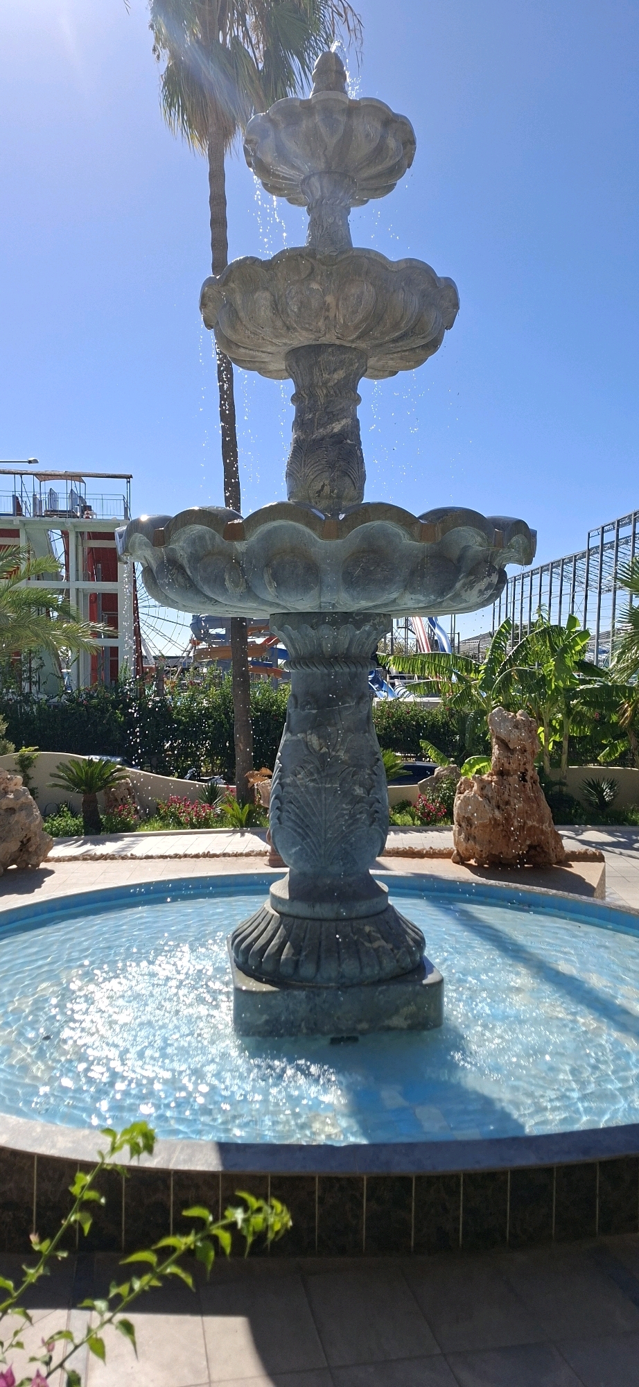 A three-tiered stone fountain with water cascading down is the central focus of the image. The fountain is situated in a bright, sunny outdoor environment with a clear blue sky. In the background, several elements suggest a resort or entertainment complex. To the left, a tall palm tree stands beside the fountain, and behind it, structures resembling water slides and scaffolding are visible. Lush green bushes and some scattered rocks, one a prominent reddish-brown boulder, frame the fountain area. The water in the fountain's basin is a vibrant blue, reflecting the sunlight. The ground around the fountain appears to be paved with light-colored tiles. In the lower left corner of the image, a leafy plant with small pink flowers is partially in focus. The scene evokes a sense of a vacation destination in Kemer, Turkey, possibly a hotel or a water park, given the presence of the water slides and resort-like structures.