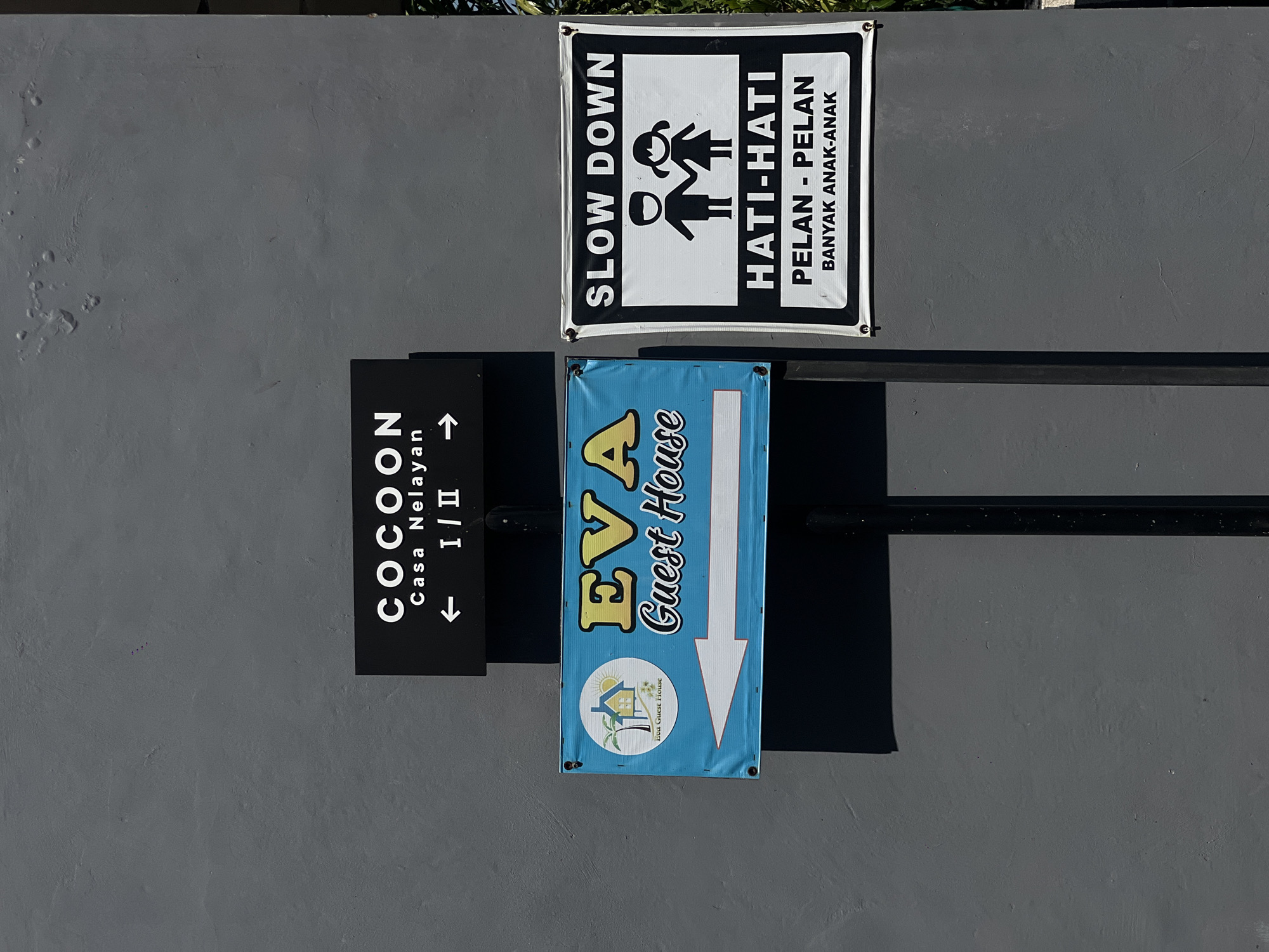 The image shows a grey wall in North Kuta, Indonesia, with three signs attached to it. At the top is a black and white sign with the text "SLOW DOWN" and "HATI-HATI PELAN - PELAN BANYAK ANAK-ANAK" alongside a graphic of two children holding hands. Below it, a blue sign with yellow lettering reads "EVA Guest House" accompanied by a downward-pointing arrow and a logo featuring a house and palm tree. To the left is a black sign displaying "COCOON" and "Casa Nelayan I/II" with left and right arrows. A black horizontal bar runs behind the signs.