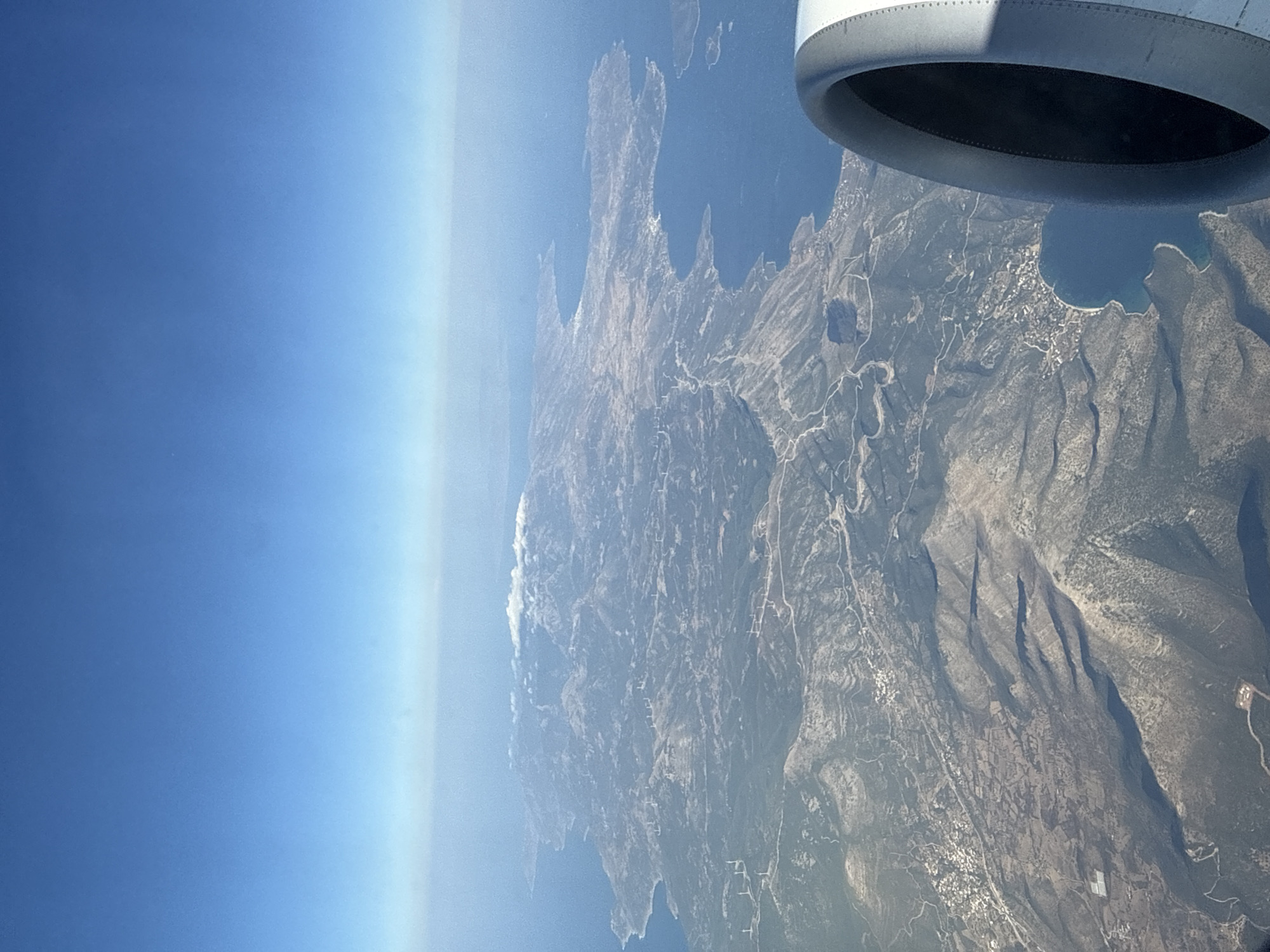 An aerial view from an airplane window shows a rugged, mountainous island coastline meeting a deep blue sea under a clear, pale blue sky. The island's terrain is a mosaic of tan, brown, and green patches, crisscrossed by winding roads and dotted with numerous wind turbines, particularly visible on the higher elevations. Small clusters of buildings, suggesting villages or towns, are scattered throughout the landscape, some nestled in coastal coves and others on the hillsides. The sea appears calm, with some small islands visible in the distance. In the foreground on the right, a portion of an airplane engine nacelle is visible, its metallic gray surface prominent against the natural scenery. The overall impression is of a remote, wind-swept island with signs of renewable energy development and scattered human settlements.