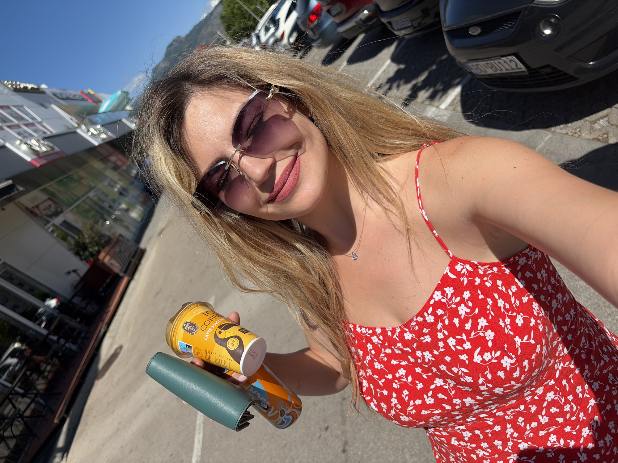A woman takes a close-up, high-angle selfie in an outdoor urban setting during a bright, sunny day. She is positioned towards the left of the frame, smiling and looking towards the camera. Her long, blonde-streaked hair flows over her shoulders, and she wears large, rimless sunglasses with tinted lenses. Her lips are painted with a soft pink lipstick, and she has a delicate silver necklace around her neck. She is wearing a vibrant red dress with a white floral pattern and thin straps.
In her left hand, she holds a yellow can labeled "ICED COFFEE" and "LATTE MACCHIATO," along with another partially obscured blue and orange can, and a dark teal/green wallet.
The background reveals a paved parking lot or street. Several cars are parked, including a black car with a visible license plate reading "BD BU112." On the left, parts of a modern building with large windows and reflective surfaces are visible, suggesting shops or commercial establishments. A clear blue sky dominates the upper part of the image, with what appears to be distant mountains in the background, partially obscured by the building. The strong sunlight casts prominent shadows, indicating midday or early afternoon. The woman's cheerful expression suggests a pleasant mood. The overall scene depicts a casual moment in a bustling area of Montenegro, specifically identified as Podgorica.
