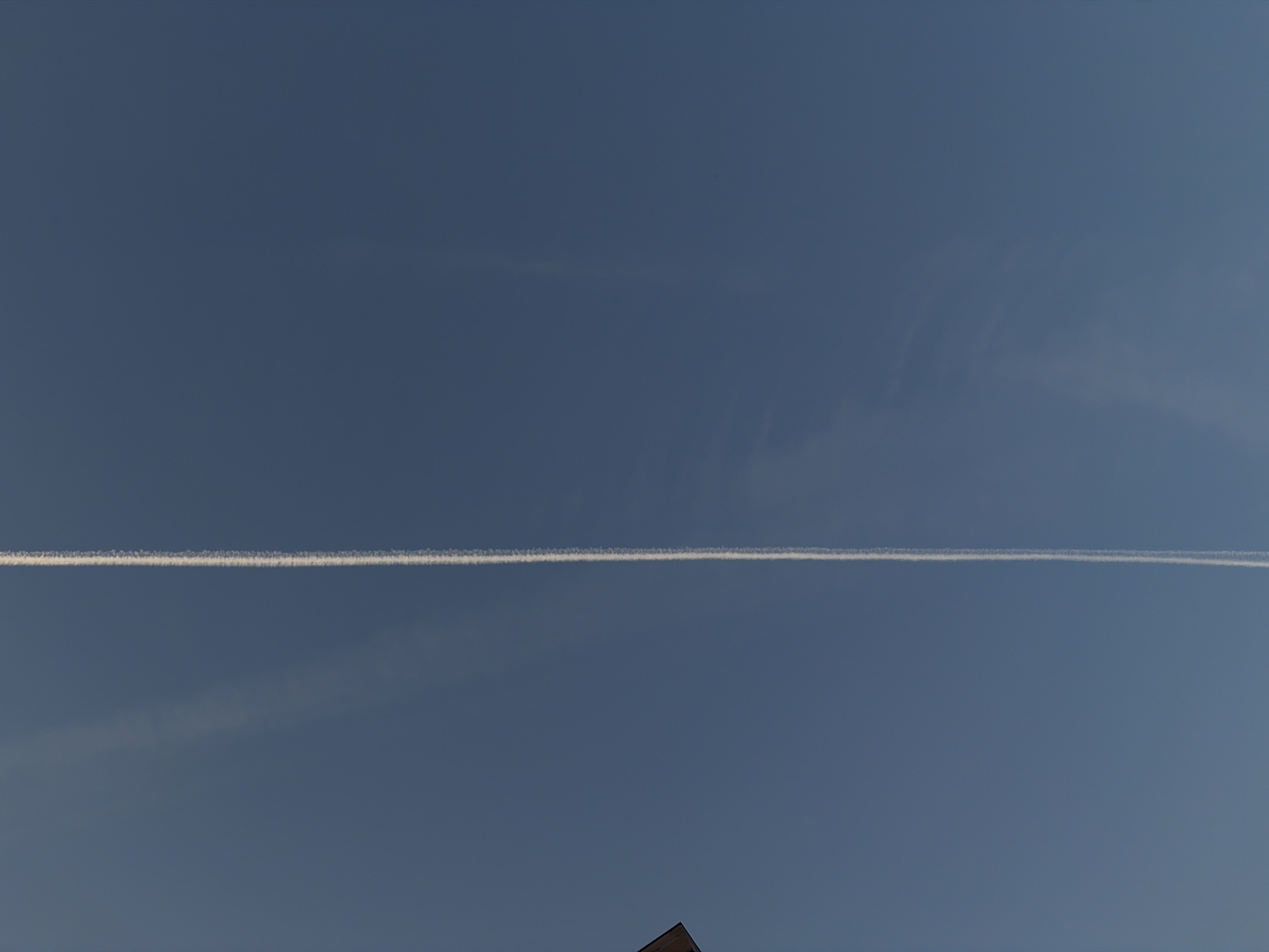 The image shows a clear blue sky with a white contrail stretching horizontally across the middle. In the lower part of the frame, a dark roof is visible. The sky is mostly uniform in color, with subtle variations in tone. The contrail appears sharp and distinct against the blue background. The location is not identifiable from the image provided.