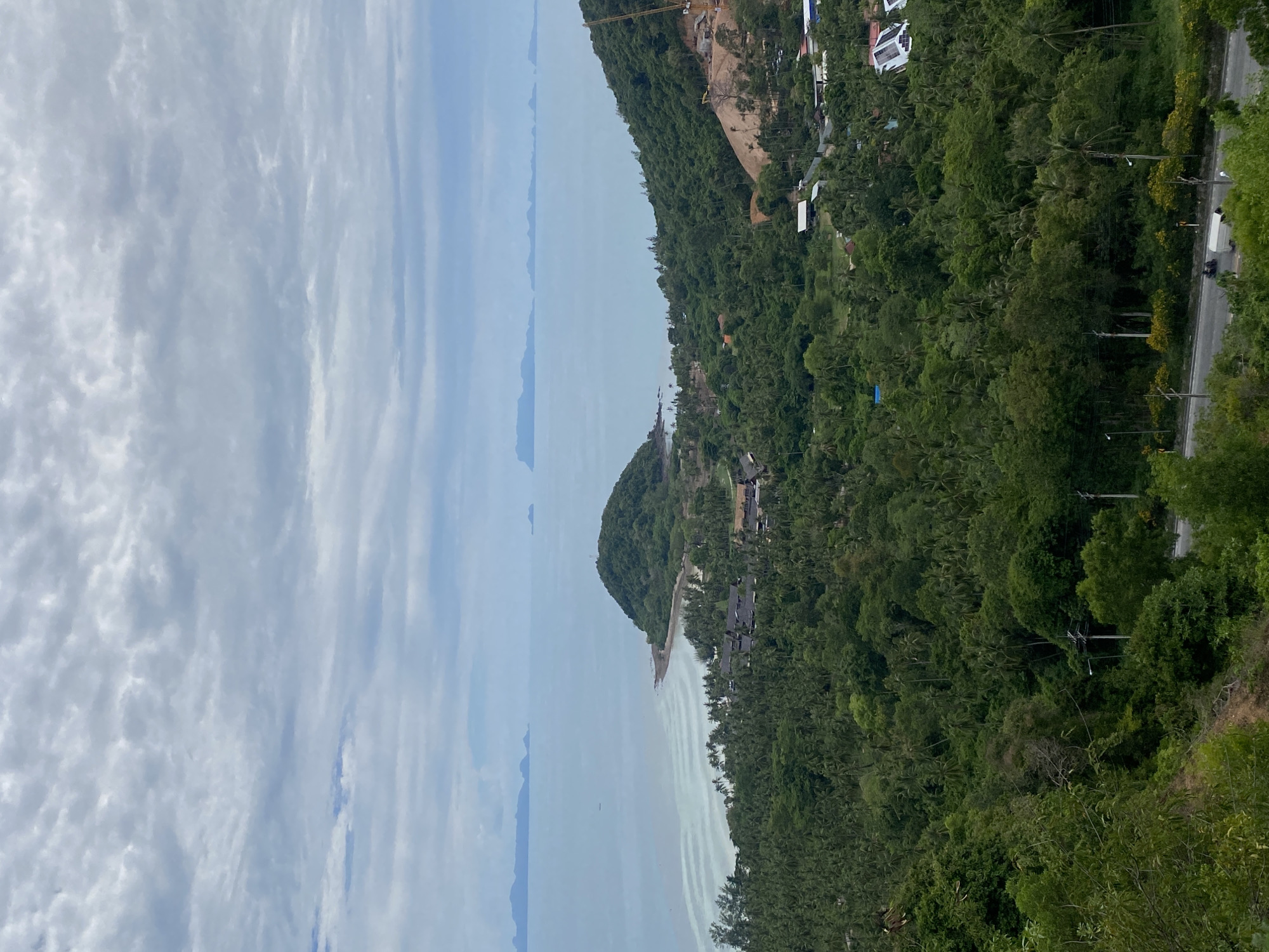 The image depicts a coastal landscape in Baan Thong Po, Thailand, dominated by lush green tropical vegetation. In the foreground, dense foliage covers a hillside, leading down to a paved road where a few vehicles, including a white van and two motorcycles, are visible. Power lines and streetlights line the road.
Further down, the landscape opens up to reveal a coastal area with a prominent, tree-covered island jutting into the turquoise sea. The island has a small settlement nestled at its base, characterized by dark-roofed buildings. The sea extends to the horizon, where several islands are faintly visible under a cloudy sky. The overall scene is one of natural beauty with signs of human development.