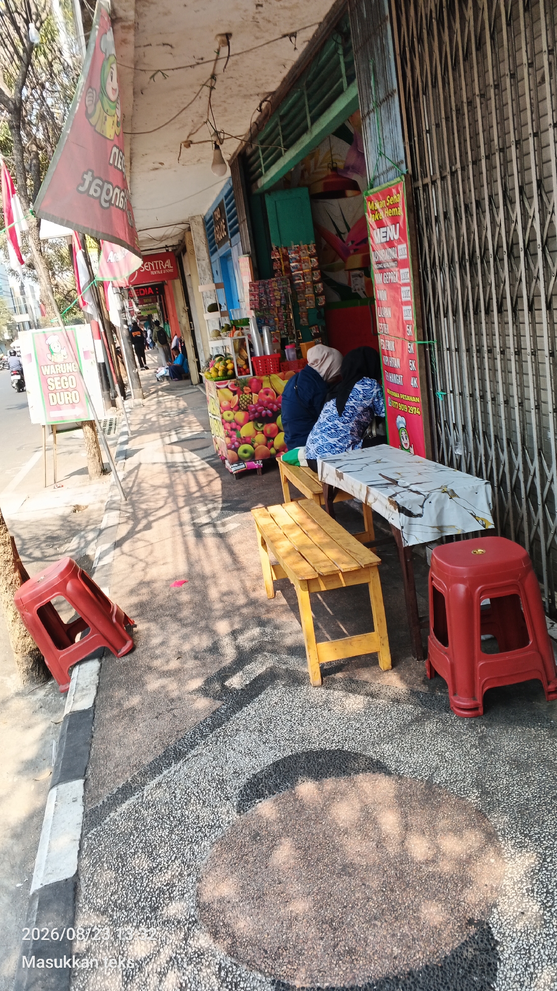 The image captures a street-level view of a food stall in Malang, Indonesia. The foreground features a red plastic stool positioned on a sidewalk with a decorative pebble pattern. To the right, a long wooden bench is paired with a table covered in a patterned cloth, where two individuals, both wearing headscarves, are seated. One person is wearing a dark blue top, and the other a blue and white patterned shirt. Adjacent to this is another shorter, yellow wooden bench. Behind them, a brightly colored display of fruits is visible, possibly part of the food stall's offerings. A menu board with Indonesian text and prices is attached to a metallic gate, which is partially covering the stall's interior. To the left, a red plastic stool is overturned. Further left, a sign for "WARUNG SEGO DURO" is displayed. The environment suggests a sunny day with dappled shadows cast by overhead trees or structures. A few other people are seen in the background walking along the street, and motorcycles are parked or passing by. The overall scene depicts a typical roadside food vendor setup in an urban Indonesian setting.