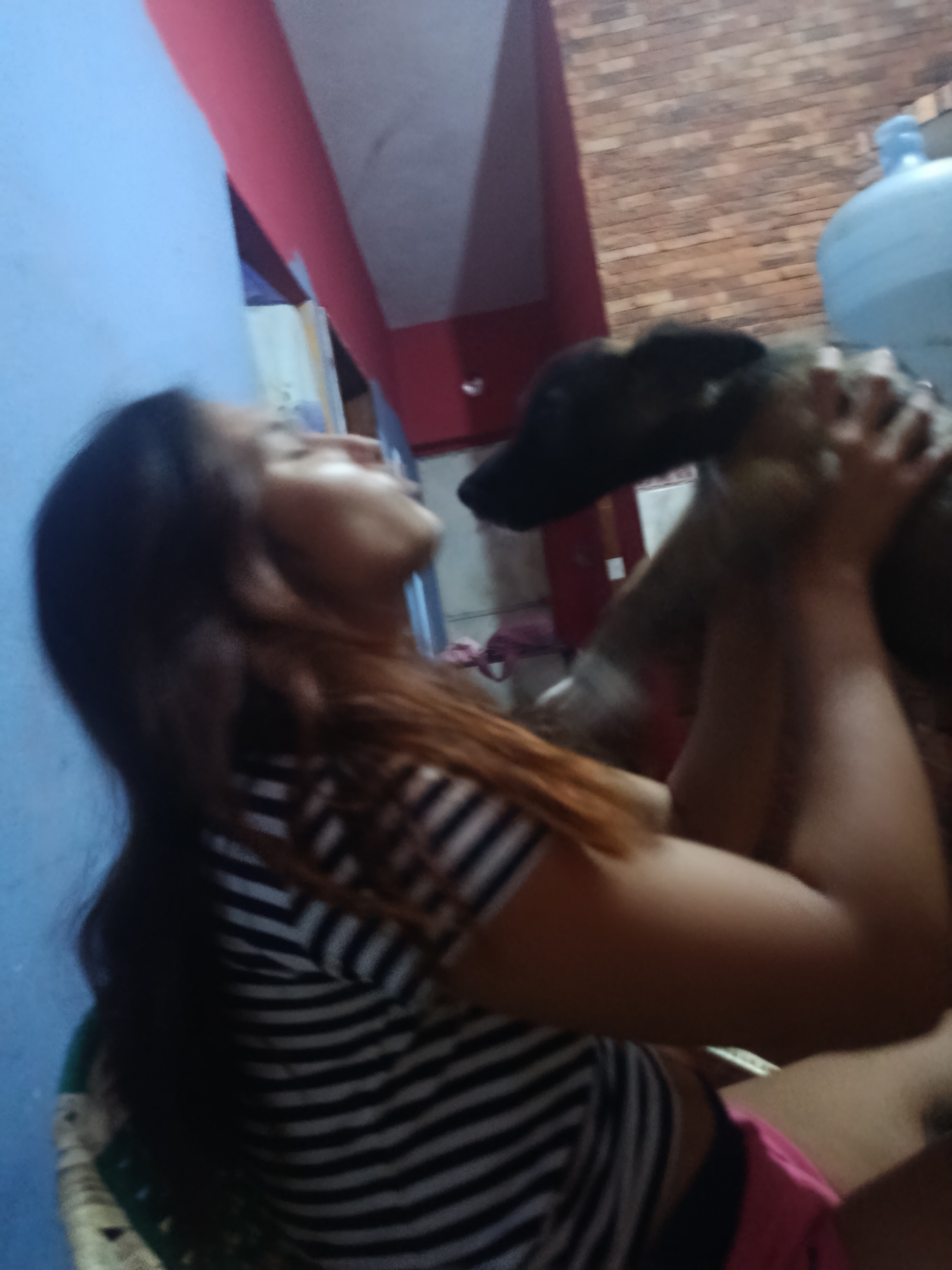 A woman with long brown hair, wearing a black and white striped shirt and pink shorts, is holding a medium-sized brindle dog. The background includes a blue wall, a brick wall, a red trim, and a large blue water jug. The location is identified as Juan Griego, Venezuela.
