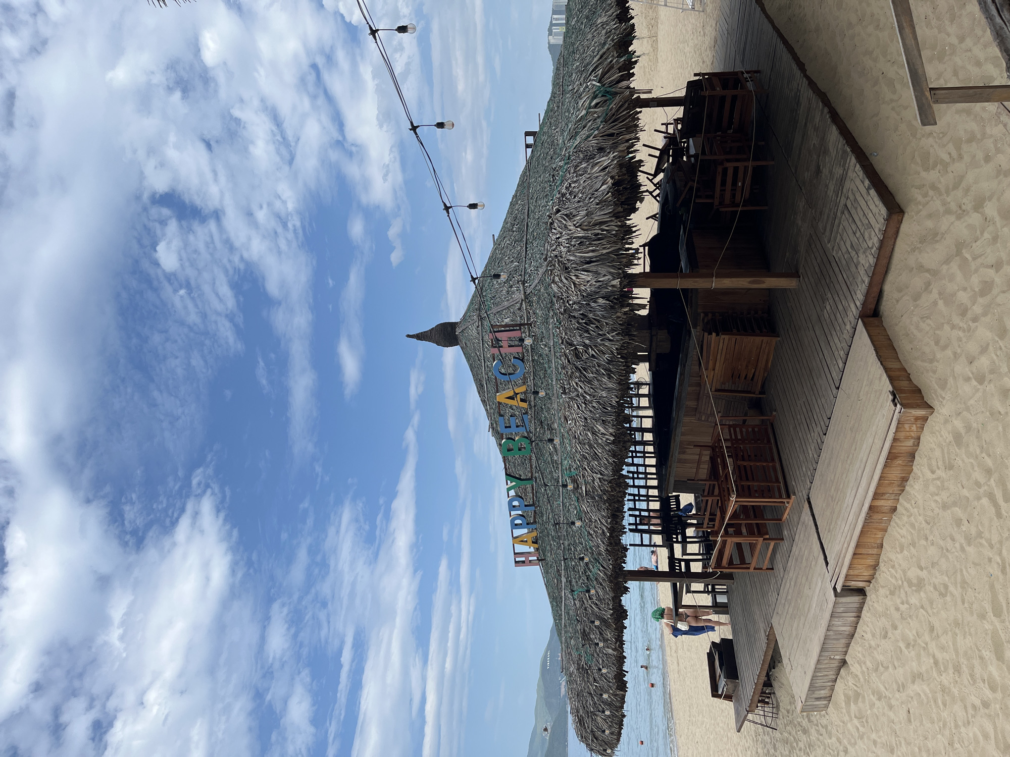 The image depicts a beach bar with a thatched roof on a sandy beach in Nha Trang, Vietnam. The bar is named "HAPPY BEACH" with colorful lettering on the roof. There are wooden tables and chairs set up on a wooden platform in front of the bar. In the background, the ocean is visible with a few people swimming. The sky is blue with some clouds. The overall scene suggests a relaxed atmosphere at a tourist destination.