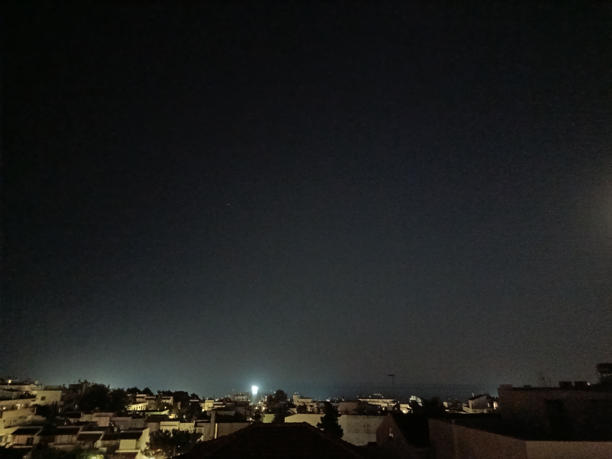 This is a night-time, high-angle, wide shot of a residential area in the Municipal Unit of Rhodes, Greece. The sky is a dark, deep blue, with a faint haze of light pollution near the horizon. The foreground is dominated by the dark silhouettes of rooftops. Further into the scene, the lights of houses and streetlights illuminate the buildings and trees of the town. A bright, concentrated light source, possibly a streetlamp or searchlight, is visible in the mid-ground, to the left of center. The overall impression is a quiet, urban night scene.