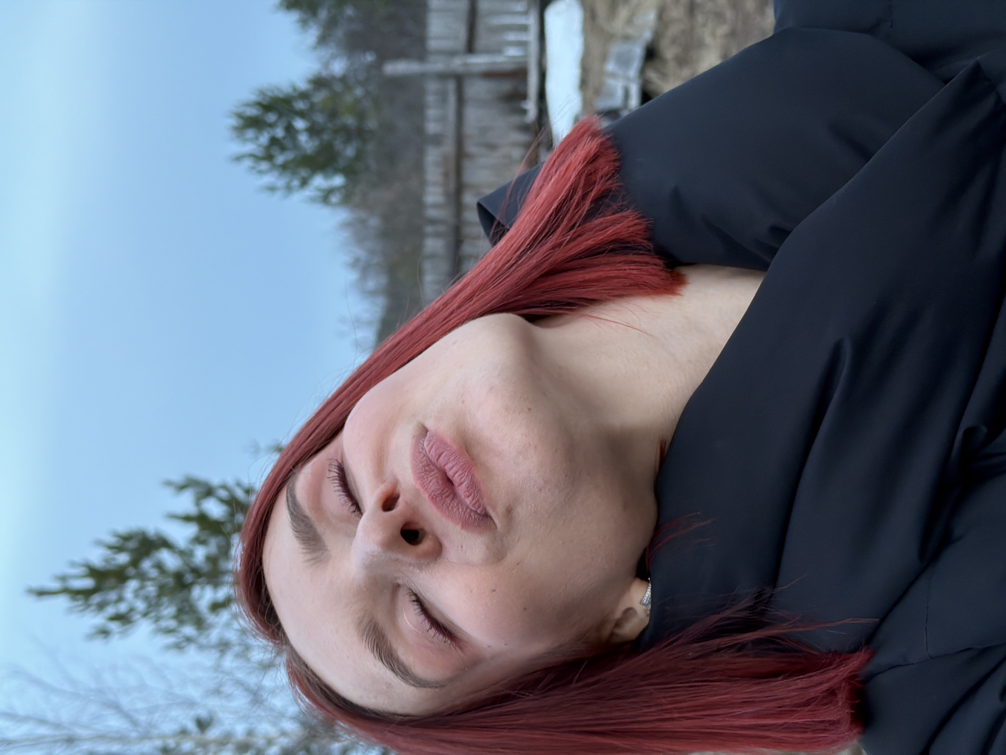A medium-shot image shows a person with red hair lying down in an outdoor setting. The person is wearing a dark, possibly black, puffy jacket. The background features a snowy landscape with bare trees and a gray sky. The location is most likely somewhere near the city of Chaikovsky, Russia, based on the prompt. The subject has their eyes closed.