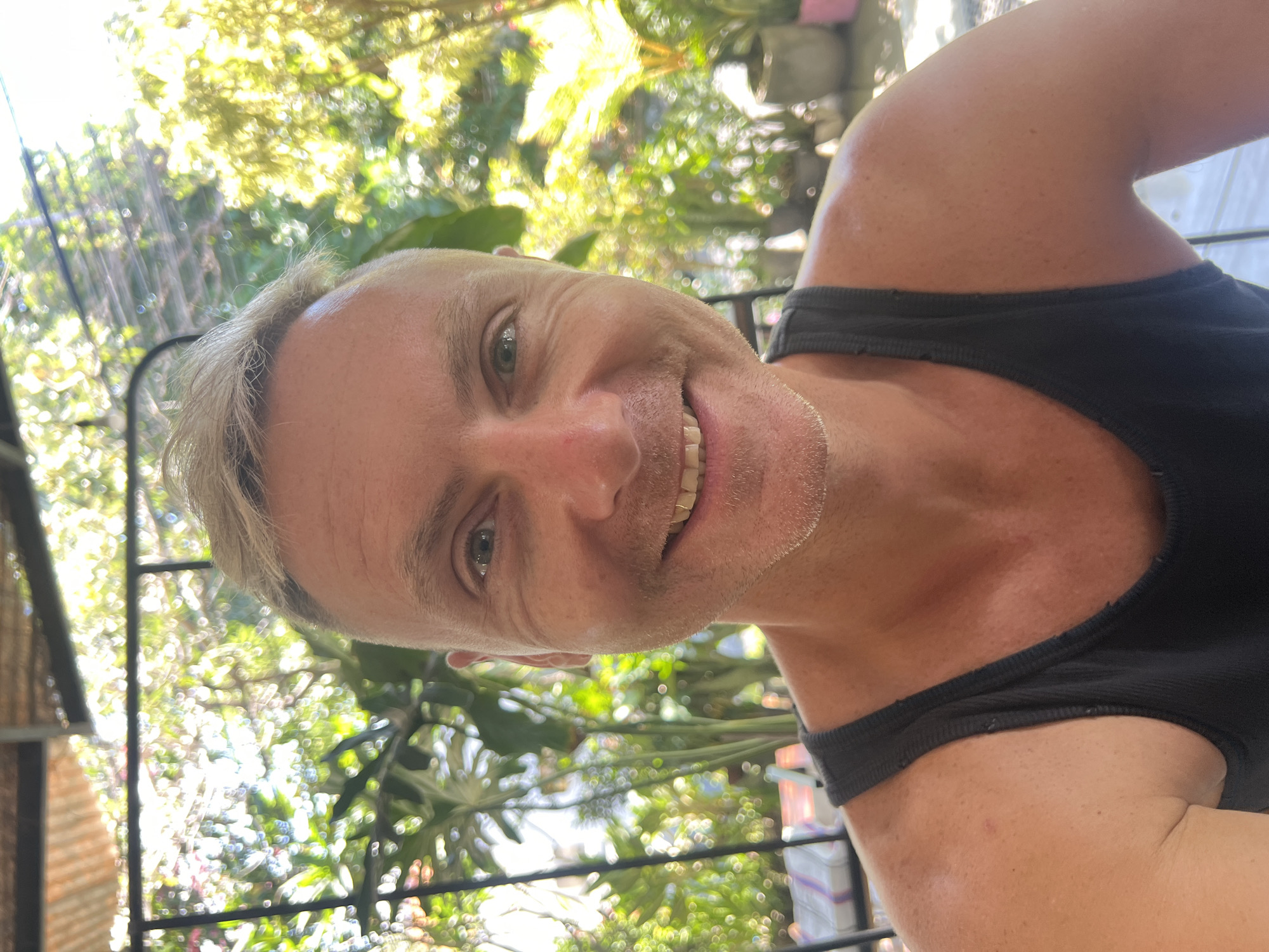 A medium shot selfie captures a Caucasian man in his late 40s or early 50s. He has short, blond hair, fair skin with some redness, and a light stubble on his face. He is wearing a black tank top. His eyes are blue and he is smiling, showing his teeth. The background is a blurred garden with lush green foliage and sunlight filtering through. The setting appears to be an outdoor patio or veranda, possibly in a tropical location, consistent with a city like North Kuta, Indonesia.