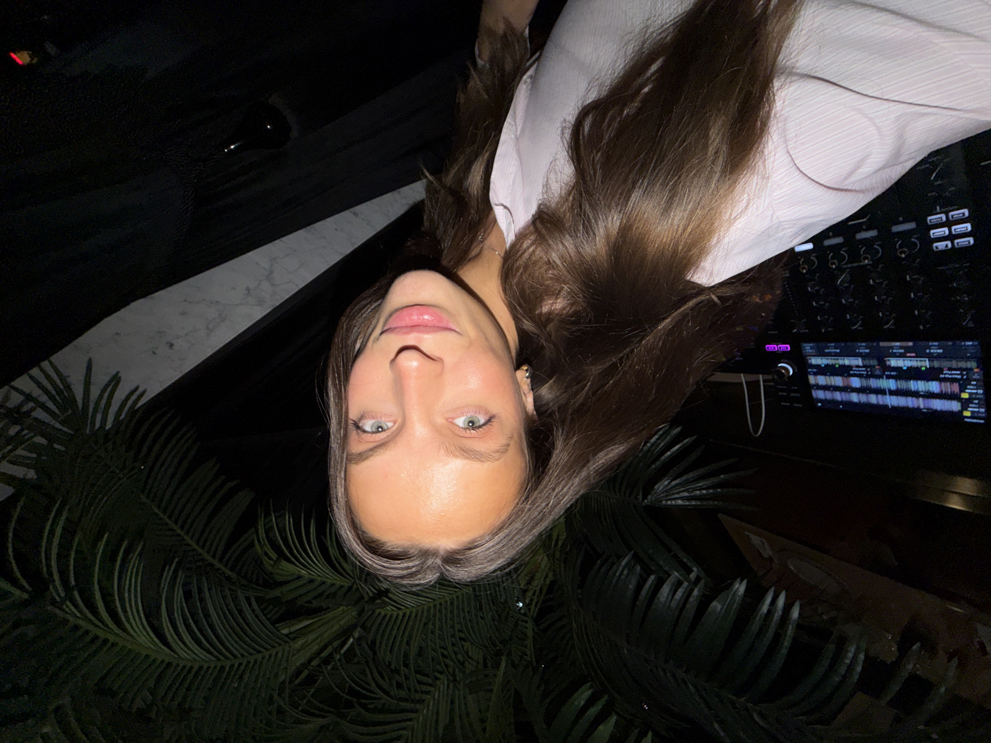 In a dimly lit environment, a young woman with long brown hair and striking blue-green eyes is captured in a selfie. She is wearing a light pink shirt with a subtle striped pattern. Her gaze is directed towards the camera, and her lips are slightly parted. She is positioned in front of lush green palm fronds, suggesting an outdoor or indoor setting with tropical plants. Behind her, to the right, a black curtain or fabric hangs, and a glimpse of a marble-patterned wall is visible. In the foreground, a black electronic device with numerous buttons and illuminated displays is partially visible, possibly a DJ console or similar equipment. The overall impression is a casual, candid moment, potentially at an event or venue in Budva, Montenegro, given the presence of tropical foliage and the sophisticated ambiance.