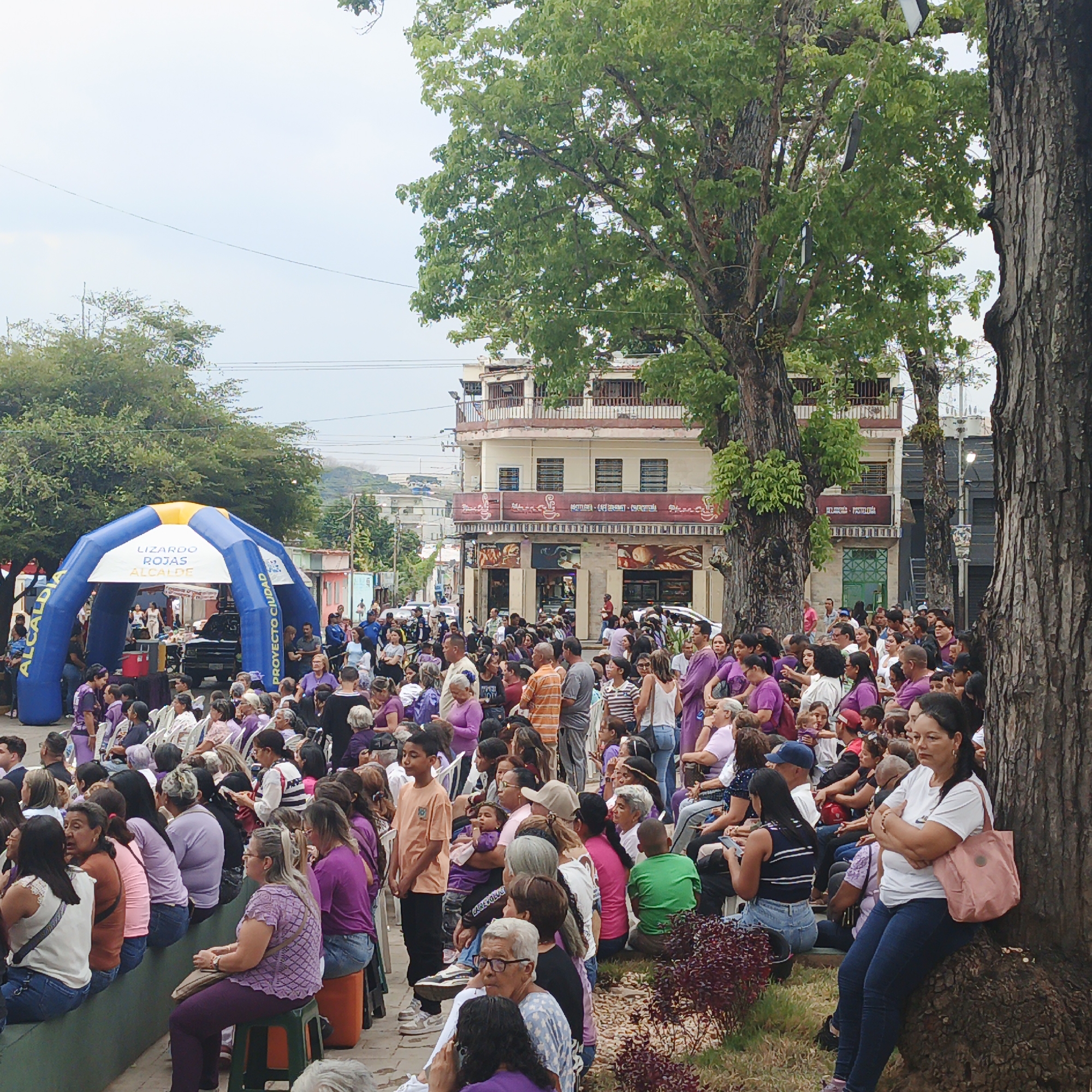 The image shows a daytime outdoor scene in Tinaquillo, Venezuela. A large crowd of people is gathered in what appears to be a city square or park. Most attendees are wearing purple clothing, possibly indicating a themed event or demonstration. Many people are seated on benches, chairs, or on the ground. An inflatable structure or arch with the words "LIZARDO ROJAS ALCALDE" is visible in the background. Buildings and trees surround the crowd.