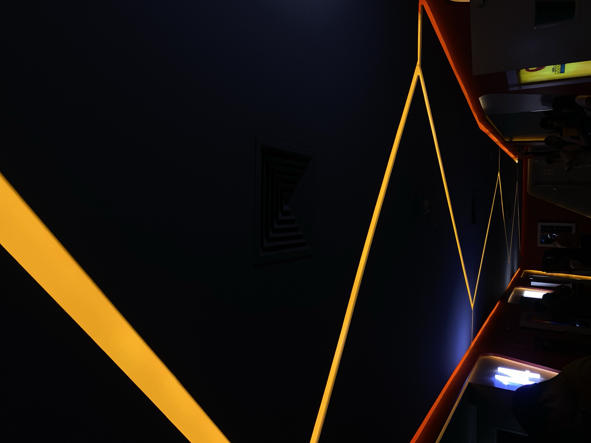 The image depicts an indoor space with a dark, moody ambiance, likely a bar or lounge in an unknown location. The dominant feature is the geometric pattern created by illuminated yellow lines against a dark blue wall, which form triangular and linear shapes. Near the upper right, a section of a counter with a red or orange trim is visible. The counter area appears to be partially occupied by blurred figures. A dark rectangular shape with lighter parallel lines is mounted on the blue wall. Additional details are obscured by shadows and the unusual perspective.