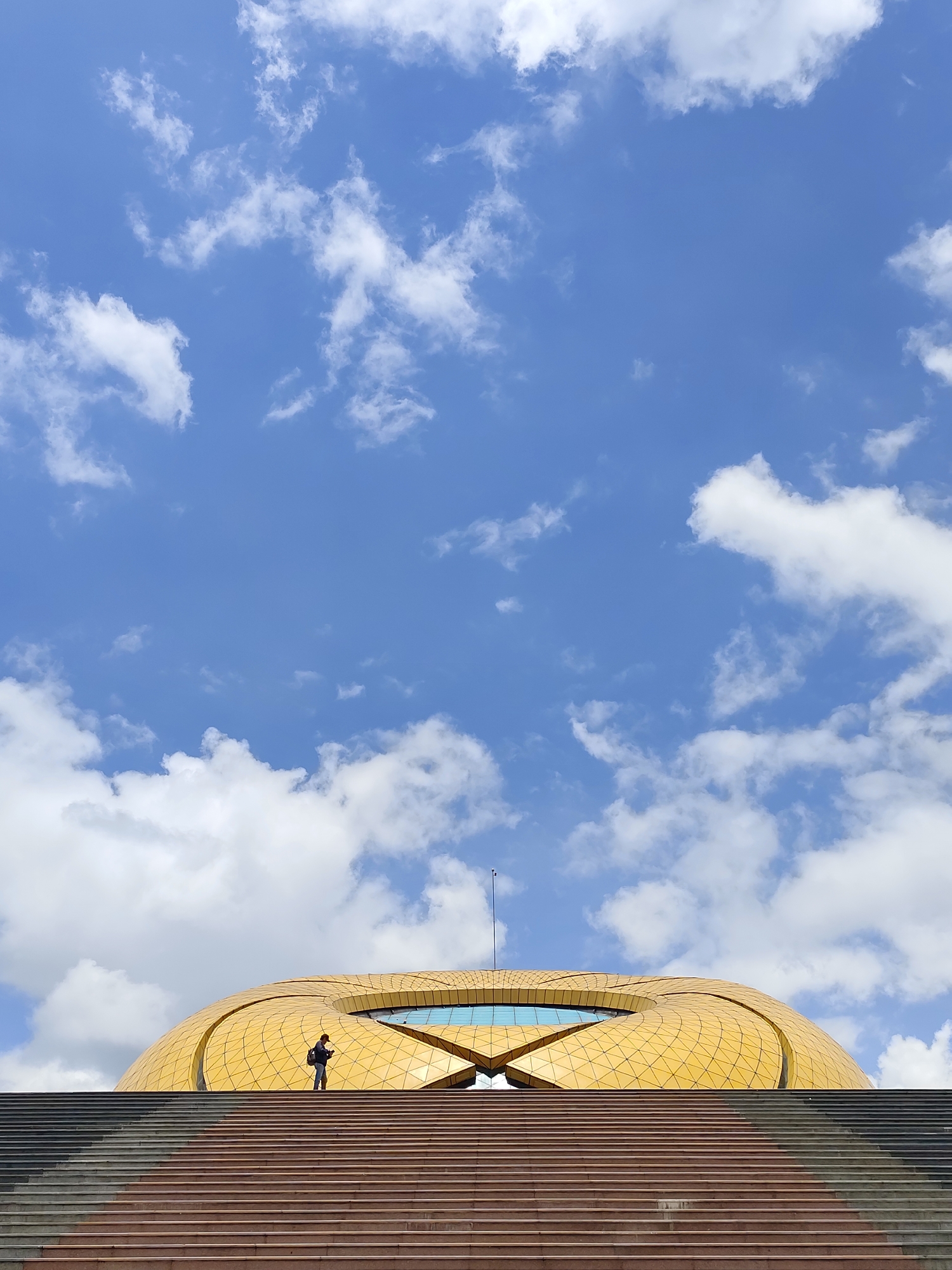 A low-angle shot presents a person standing halfway up a grand set of stairs, with a large, golden, lotus-shaped building looming behind them and a vibrant blue sky filled with puffy white clouds serving as the backdrop. The building is characterized by its golden color, lotus-like shape, triangular patterns, and glass features at the top. The staircase is divided into different segments, with some steps in a light brown color and others in a darker gray hue. A person wearing a dark jacket and carrying a backpack is seen halfway up the stairs, adding a sense of scale and human presence to the scene. A thin, vertical antenna-like structure extends from the top of the building toward the sky.