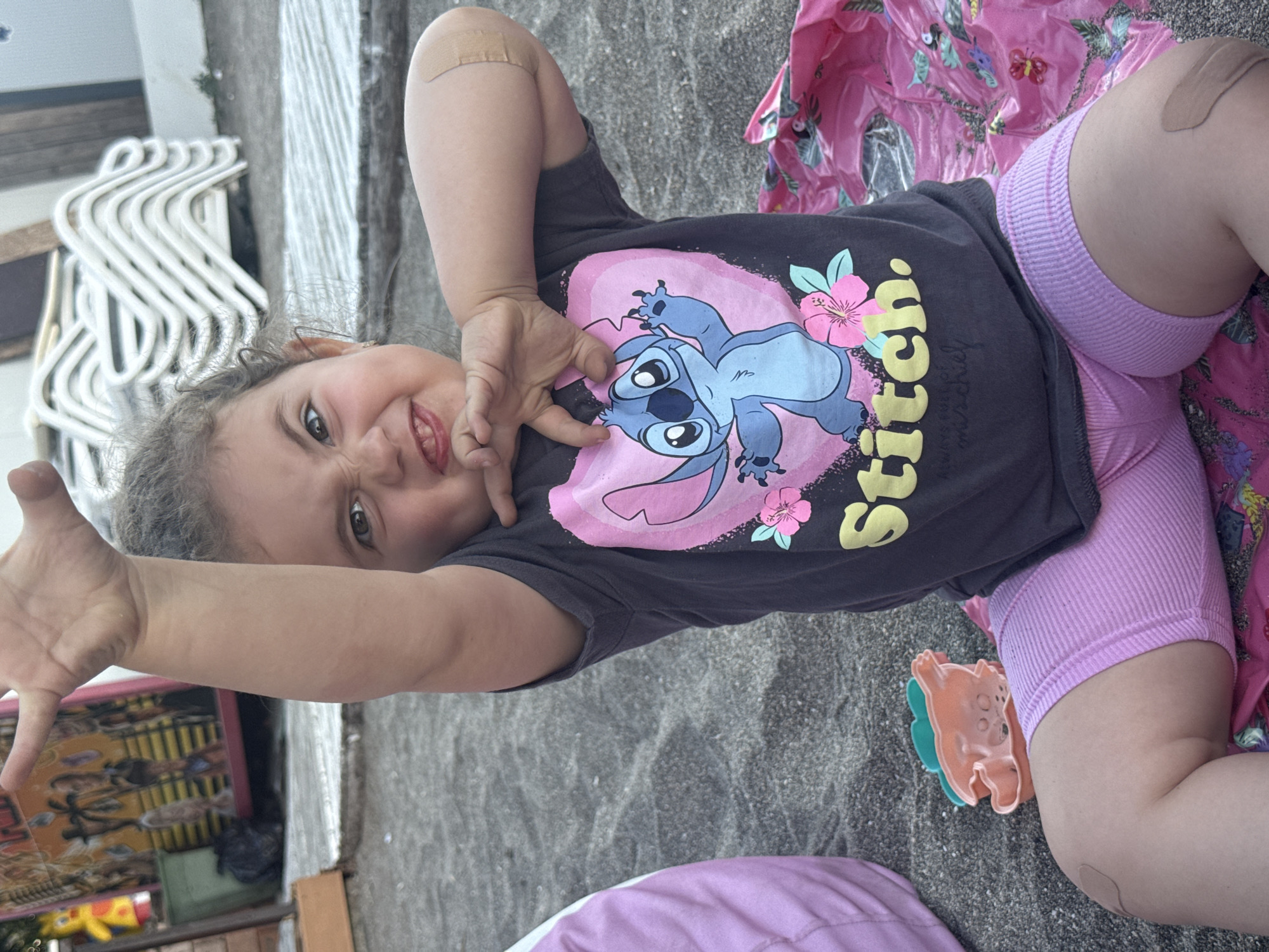 A young girl is lying on a sandy surface, possibly a beach, in Burgas, Bulgaria. She is wearing a dark t-shirt with a cartoon character, likely Stitch from Lilo & Stitch, printed on the front, along with pink shorts. A bandage is visible on her upper arm. The girl is looking towards the camera with a playful expression and has her right arm extended. Next to her, there's a pink blanket with a floral pattern and a colorful toy. In the background, rows of white lounge chairs and some vegetation are visible. The lighting suggests it could be daytime.